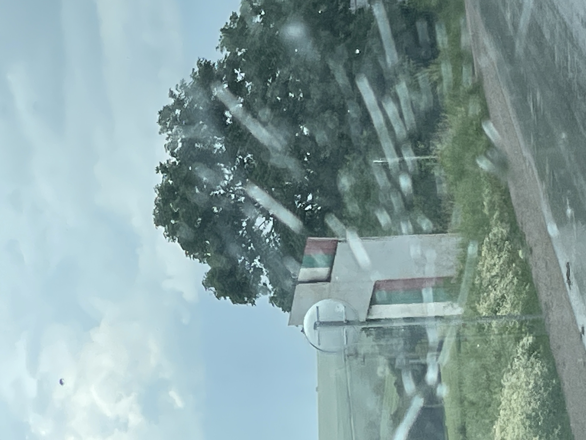 The image shows a view through a wet window. Outside, there is a building with a light gray facade and horizontal red and green stripes near the top. A tree with dense green foliage is partially obscuring the building. To the right, there's greenery, possibly bushes or grass, alongside a concrete curb or edge. The sky is visible in the upper left, with light clouds. It is not possible to determine if this image is in the city of Loznitsa, Bulgaria.
