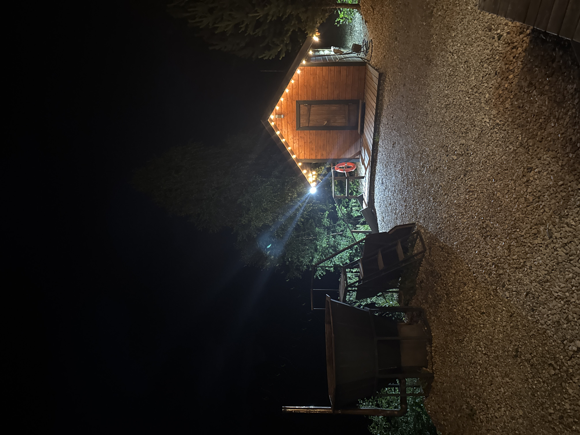 This is a nighttime, eye-level, exterior shot of a small wooden building situated on a gravel surface. The building, which appears to be a sauna or bathhouse, is illuminated by a string of warm-toned lights draped along its eaves. A wooden door is visible on the front of the structure, and a small red life preserver is hanging on the railing to its left. To the left of the building, there is a large metal tub with a chimney or pipe extending upwards, and a metal staircase or ladder structure. The scene is bordered by trees and foliage, suggesting a rural or natural setting. The overall atmosphere is dimly lit and somewhat rustic. This image was taken in Urazovo, Russia.