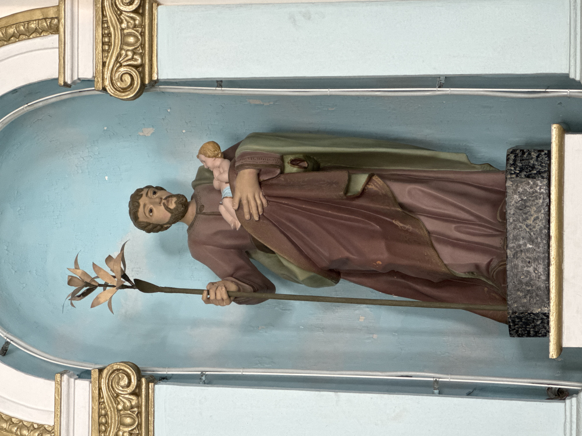 The image shows a statue of Saint Joseph holding the infant Jesus. Saint Joseph is depicted with a beard and is wearing a reddish-brown robe and a green cloak. He is holding a staff with a metallic flower-like decoration in his left hand. The infant Jesus is being held in his right arm. The statue is positioned in an alcove with a light blue, textured background. The alcove is flanked by decorative golden columns. The statue stands on a black and grey speckled base. The scene appears to be inside a church or religious building in Niterói, Brazil.