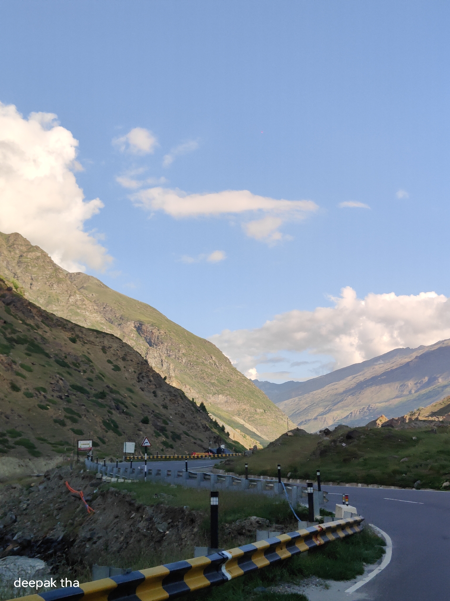 The image depicts a winding mountain road in Sissu, India, with steep, verdant slopes on either side and a clear blue sky with scattered clouds overhead. The road is bordered by a yellow and black guardrail, and a few traffic signs are visible. The terrain suggests a high-altitude location, likely in the Himalayas. The image is a photograph taken in daylight, capturing the grandeur of the natural landscape. There are no people visible in the frame. The overall scene conveys a sense of travel and exploration through a remote mountain region.