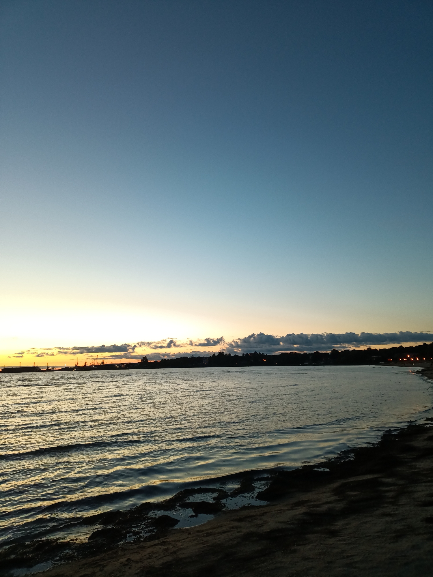 The image depicts a tranquil seascape at dusk, likely overlooking the Tallinn Bay in Tallinn, Estonia. The sky transitions from a deep blue at the top to a pale yellow and orange hue near the horizon, indicating the aftermath of sunset. A line of dark clouds is visible along the horizon, partially obscuring the distant shoreline.
On the left side of the horizon, a silhouette of industrial structures, possibly cranes and buildings of a port, is visible against the bright sky. The water in the foreground is calm, with gentle ripples reflecting the fading light. The shoreline on the right is dark and textured, with some rocks or debris visible near the water's edge. A few scattered lights are discernible along the distant coastline, suggesting the presence of a town or settlement. No people or specific actions are identifiable in the scene. The overall atmosphere is serene and peaceful, capturing the quiet beauty of the transition from day to night over the water.