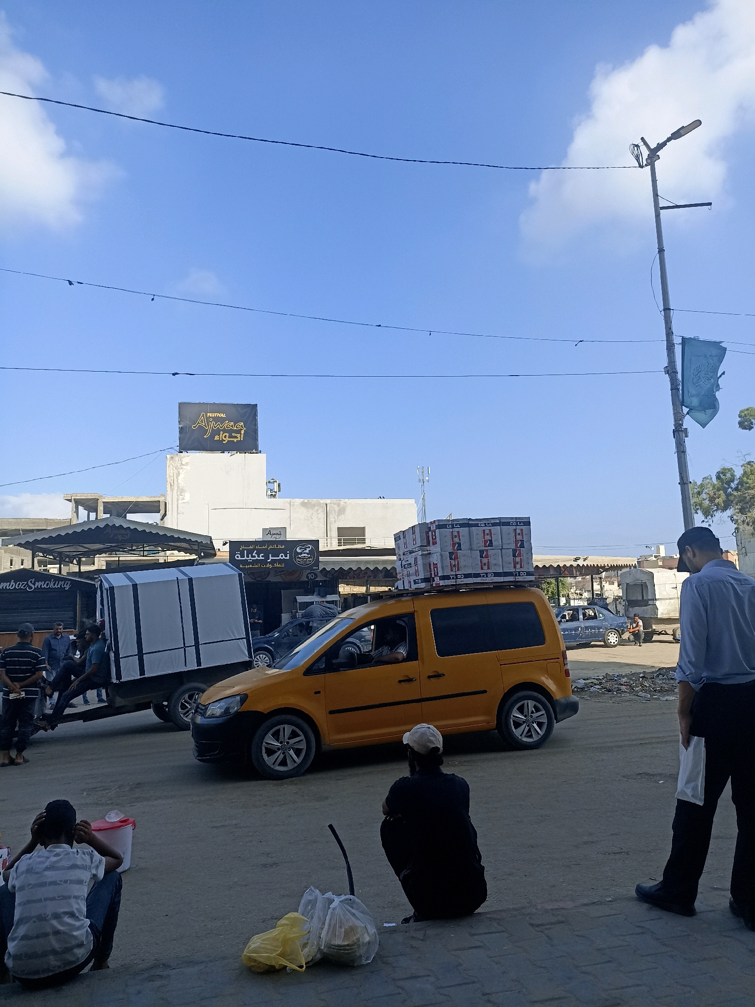 A street scene in the city of Gaza, Palestinian Territories, shows an outdoor environment with a clear blue sky and some clouds. In the foreground, several people are seated on the ground, some looking away from the camera. A yellow van is parked on the street, loaded with boxes on its roof. A white truck with a covered trailer is parked to the left of the van. In the background, buildings and a lamppost are visible. Several signs are displayed, including one with Arabic text and another that says "FESTIVAL Ajwaa." Debris is scattered on the ground. A person in a blue shirt and black pants stands on the right, facing away from the camera, holding a white bag. The overall impression is a busy street with commercial activity and pedestrian presence.