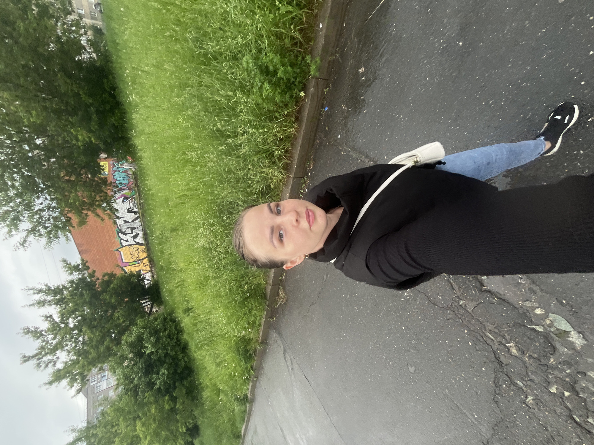 A young woman stands on a cracked asphalt road next to a curb. To her left is tall green grass and a brick building with graffiti. She wears a black hooded jacket, blue jeans, and black and white sneakers. She has a white bag over her shoulder. The background shows a city building through the trees. This location is in Izhevsk, Russia.