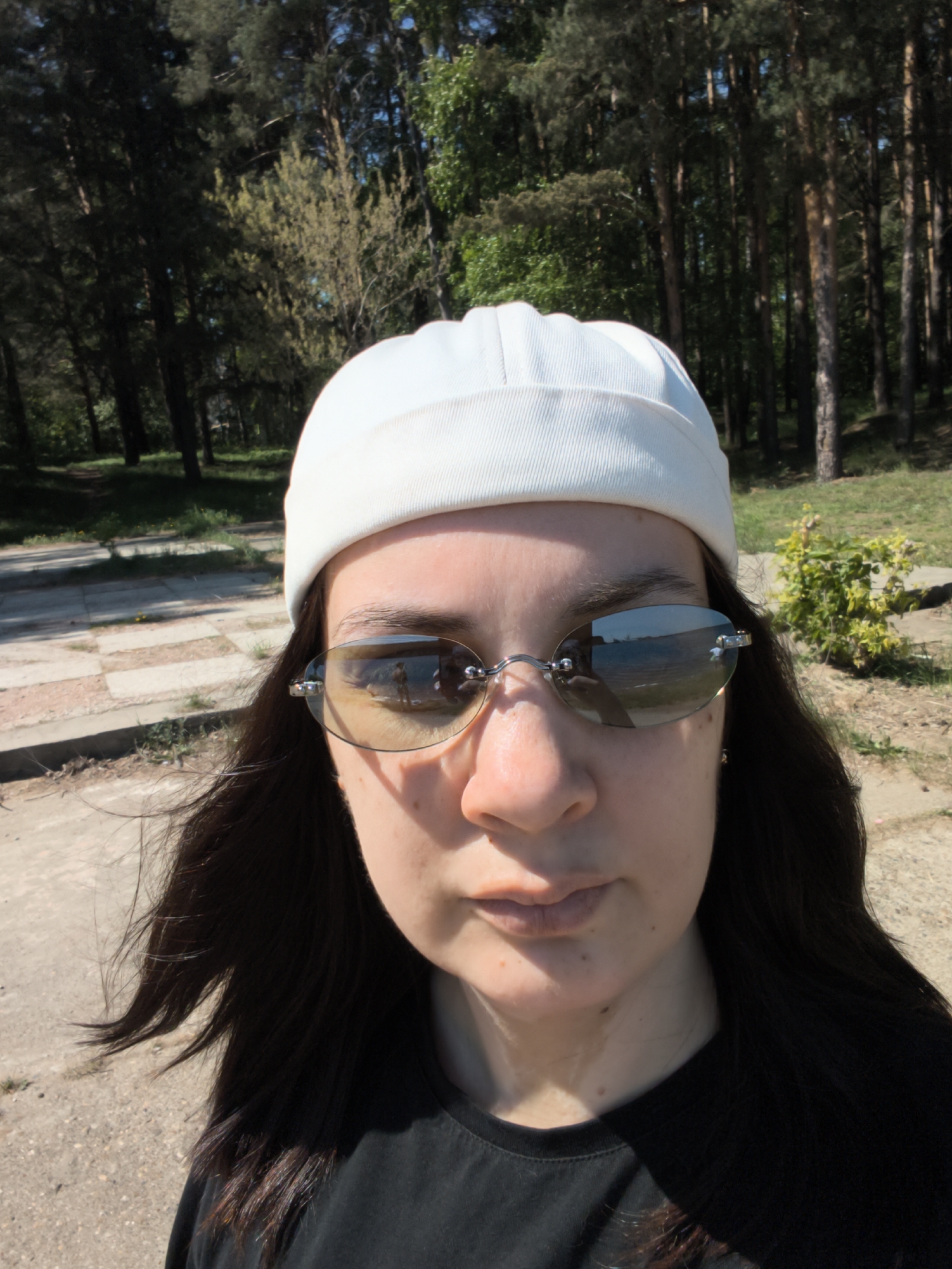 A close-up selfie shows a person with dark hair, wearing a white beanie and frameless sunglasses. The sunglasses reflect a scene with water, trees, and figures. The person is wearing a black t-shirt. The background features trees and greenery, suggesting a park or wooded area. A paved path is visible to the left. Based on the presence of pine trees and the general setting, the image may have been taken in or near a park in Chaikovsky, Russia.