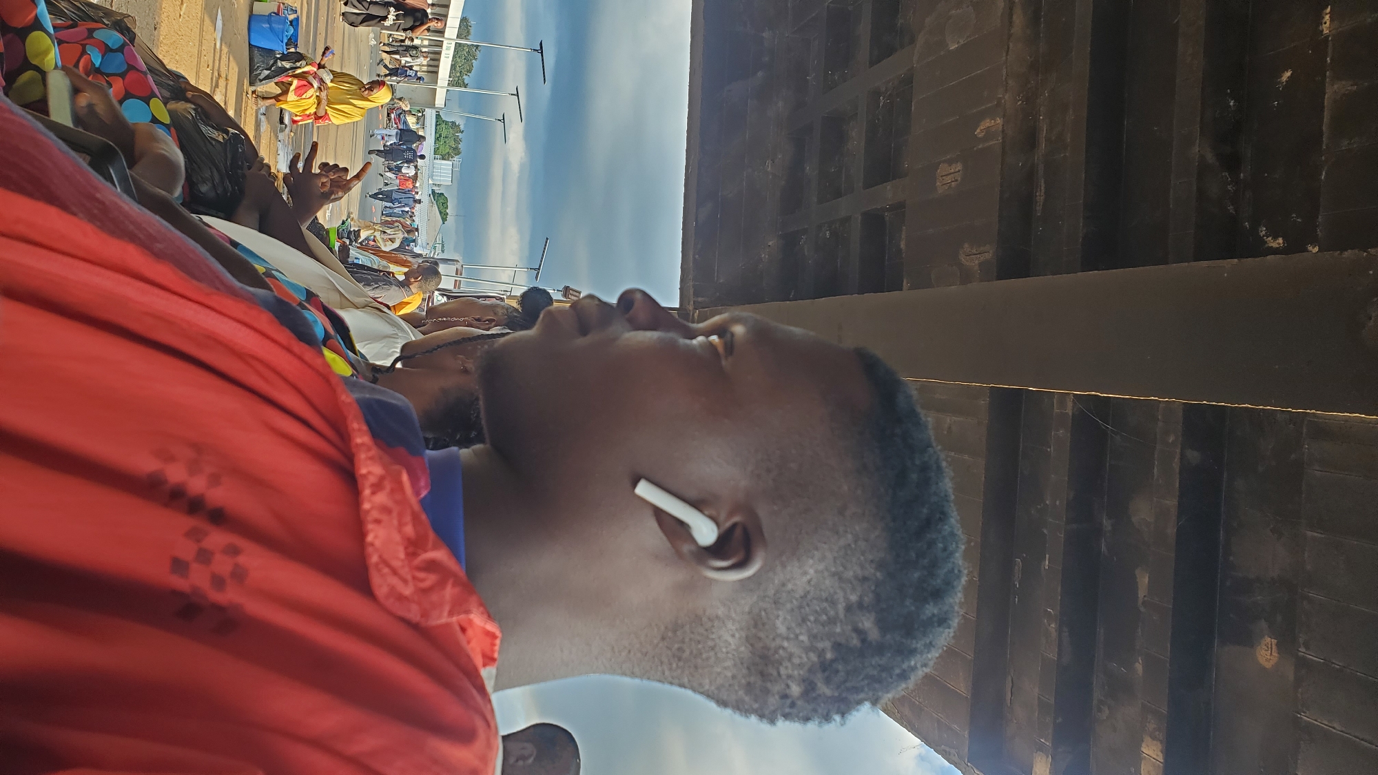 A side profile of a young Black male is captured, facing left, in the foreground. He has a short haircut and is wearing a white earbud in his right ear. His attire consists of a bright orange jacket over a blue shirt. Behind him, partially visible, is the side of another individual, also in the foreground, wearing a colorful patterned garment.
The background suggests an outdoor, likely urban, setting. A large, dark, concrete structure, possibly a bridge or an overpass, dominates the upper portion of the image, casting a shadow. Below this structure, a bustling scene unfolds. A group of people are gathered, with some sitting and others standing. To the left, a woman in a yellow headscarf and robe is seated next to a blue bucket. Further back, a crowd of people walks in the distance, with buildings and streetlights visible. The sky is overcast with dramatic, dark clouds. The ground appears to be dirt or unpaved. The overall impression is of a public space, possibly a market or a transit area, under a large overhead structure.