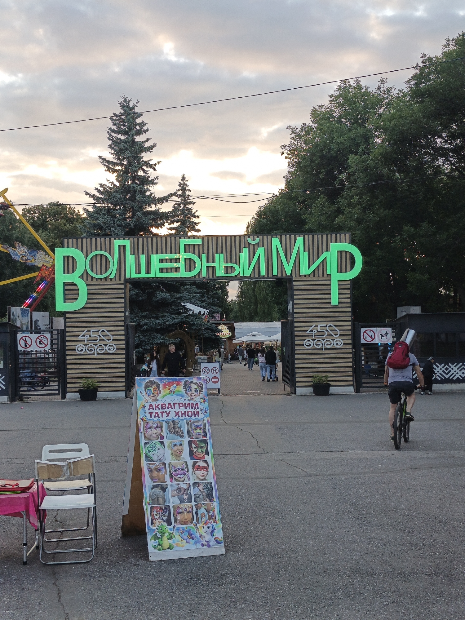 The image depicts an outdoor scene, likely an amusement park or entertainment venue, in Ufa, Russia. A large, brightly lit green sign with Cyrillic text reads "ВОЛШЕБНЫЙ МИР" (Volshébnïy Mir), meaning "Magic World." The sign is mounted above an arched entryway, supported by wooden pillars adorned with circular decorative elements.
In the foreground, a man rides a bicycle away from the camera, wearing a red backpack. To the left, a small table covered with a pink cloth displays some items, and two white folding chairs are placed nearby. A prominent A-frame sign stands to the right of the chairs, showcasing a colorful display of face paint designs for children, including animals, superheroes, and fantasy characters, with the text "АКВАГРИМ ТАТУ ХНОЙ" (Akvagrim Tatu Khnoy) meaning "Face painting tattoo henna."
The background shows a pathway leading into the park, with trees lining the sides. Several people are seen walking through the entrance. A Ferris wheel-like amusement ride is partially visible on the far left. The sky is cloudy, suggesting it might be late afternoon or early evening. The overall atmosphere appears lively and recreational, characteristic of a public entertainment space.