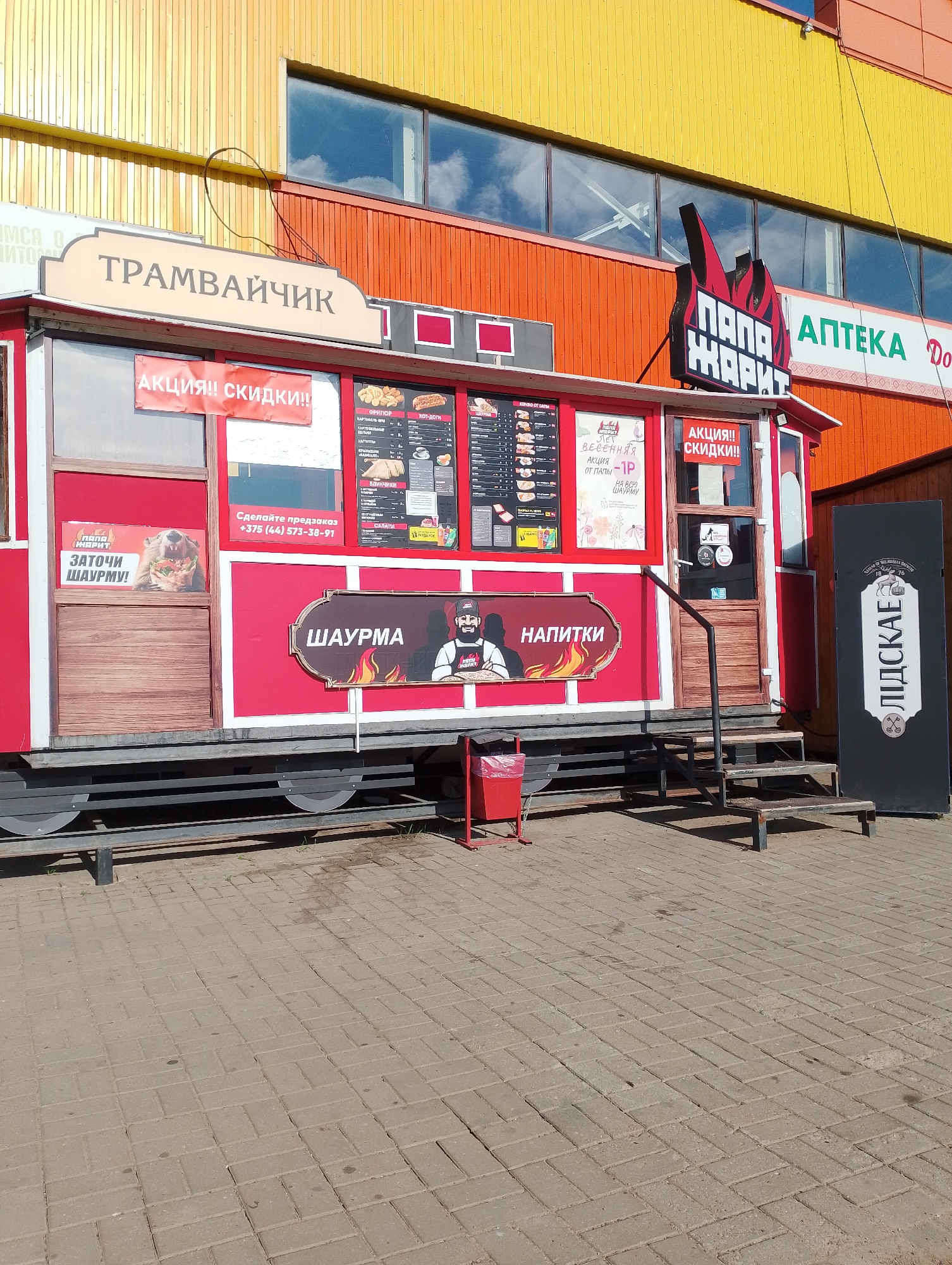 The image shows a red food trailer, painted to resemble a tram, with various signage and advertisements. The trailer is labeled "ТРАМВАЙЧИК" (Tramvaychik) on the top. To the right, a sign with flames reads "ПАПА ЖАРИТ" (Papa Zarit), which translates to "Papa Grills." There are also signs for "АПТЕКА" (Apteka - Pharmacy). On the trailer itself, there are advertisements for "АКЦИЯ!! СКИДКИ!!" (Action!! Discounts!!), and a menu board with items like "ШАУРМА" (Shawarma) and "НАПИТКИ" (Drinks). A sign displays a bear holding a shawarma and the text "ЗАТОЧИ ШАУРМУ!" (Sharpen your shawarma!). The trailer is parked on a paved area, with a set of metal stairs leading up to the entrance. A red trash can is visible near the stairs. In the background, a building with yellow and orange facades and windows can be seen. The overall scene appears to be an outdoor food vendor located in a commercial area, likely in Vitsebsk, Belarus, given the Cyrillic script and the mention of a pharmacy. There are no people clearly visible in the image.