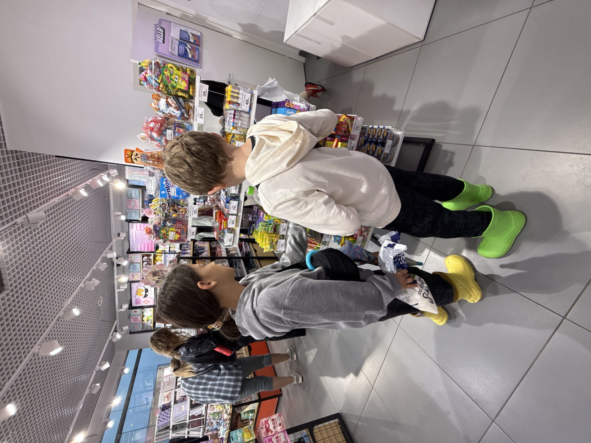 The image depicts the interior of a retail store, likely a gift shop or convenience store, located in Nizhny Novgorod, Russia. The store features a brightly lit ceiling with a grid of fluorescent lights and a white drop ceiling. Shelving units are visible, stocked with various merchandise, including packaged snacks, candies, and possibly novelty items. The flooring is a light gray tile with a grid pattern.
In the foreground, two children are observing the merchandise. A girl with dark hair, wearing a gray hooded sweatshirt and dark pants, stands on the left. She is wearing bright yellow rain boots and holding a white, crinkled plastic bag. To her right, a boy with light brown hair, wearing a light cream-colored hooded sweatshirt and dark pants, is also looking at the shelves. He is wearing lime green rain boots.
Behind the children, other shoppers are present. A woman with curly brown hair, wearing a black jacket over a plaid shirt, is in the middle ground. To her left, another person, partially obscured, is wearing a gray and white plaid shirt and gray pants.
The merchandise on the shelves includes colorful packaging, with some price tags visible in Russian currency. For instance, price tags such as "39.", "79.", and "49." are discernible. The overall atmosphere suggests a commercial environment where items are displayed for purchase.
