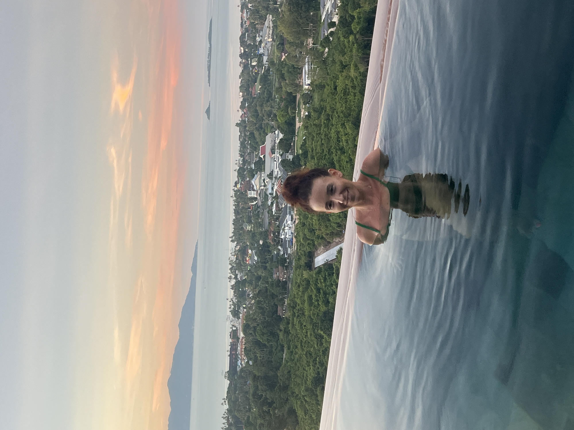 A woman with dark hair pulled into a bun and wearing a green bikini top is standing in an infinity pool. She is smiling and looking at the camera. The pool overlooks a lush green landscape with buildings and palm trees, leading to the ocean and distant islands. The sky is a soft gradient of orange, pink, and blue, indicating it is either sunrise or sunset. The image appears to have been taken in Baan Bo Phut, Thailand, given the coastal scenery and tropical vegetation.