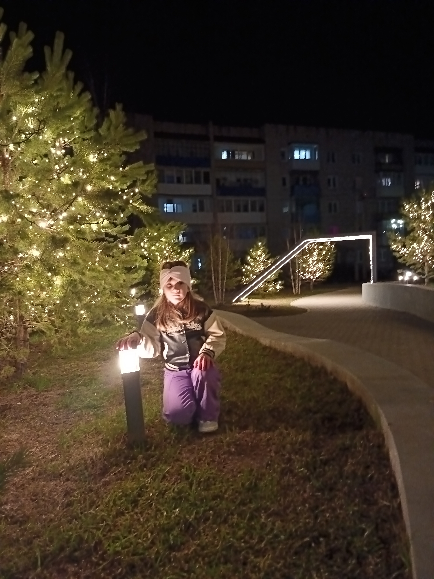 A young girl is kneeling on a patch of grass in Osa, Russia. The girl has fair skin, brown hair, and is wearing a white headband. She is wearing a letterman jacket and purple sweatpants. Her right hand rests on a small, black lamp post that is illuminated. Behind her are several pine trees decorated with string lights. In the background is a multi-story building with several lighted windows. A sidewalk runs behind the girl, and there is a large, rectangular lighting structure over the sidewalk.