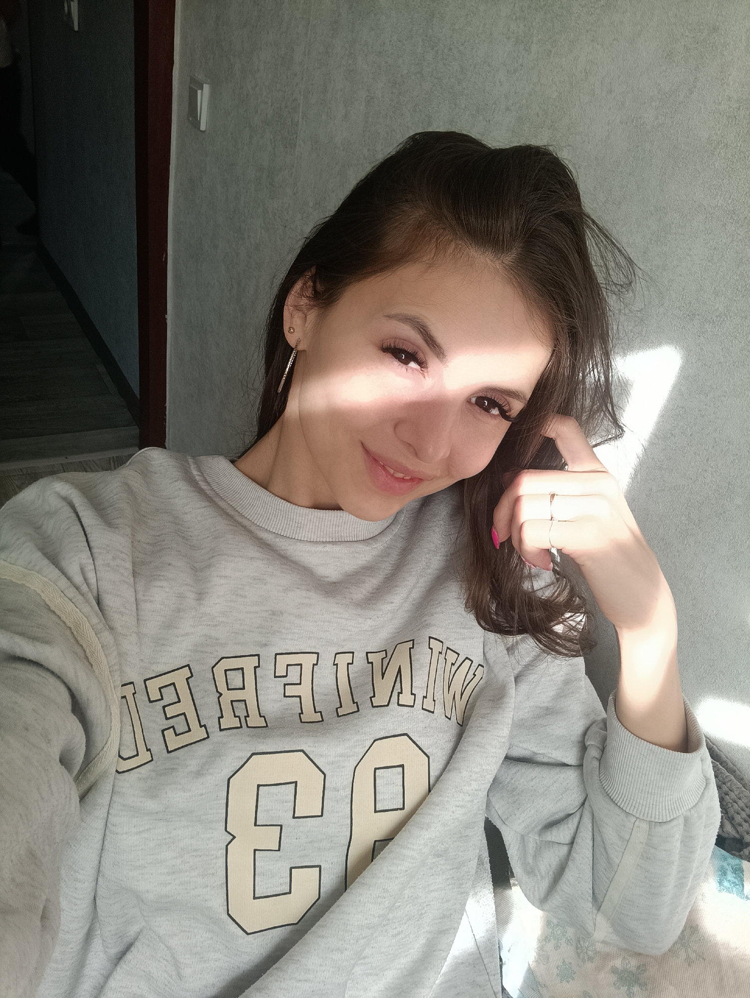 A woman is sitting in an indoor location with a light-grey wall. She has long, dark hair and is wearing a grey sweatshirt with the word "WINNIPEG" in mirrored text and the number "93" on it. She is smiling and holding her hand up near her face. Sunlight is shining on her face and the wall. The location is in the city of Unknown, Unknown.
