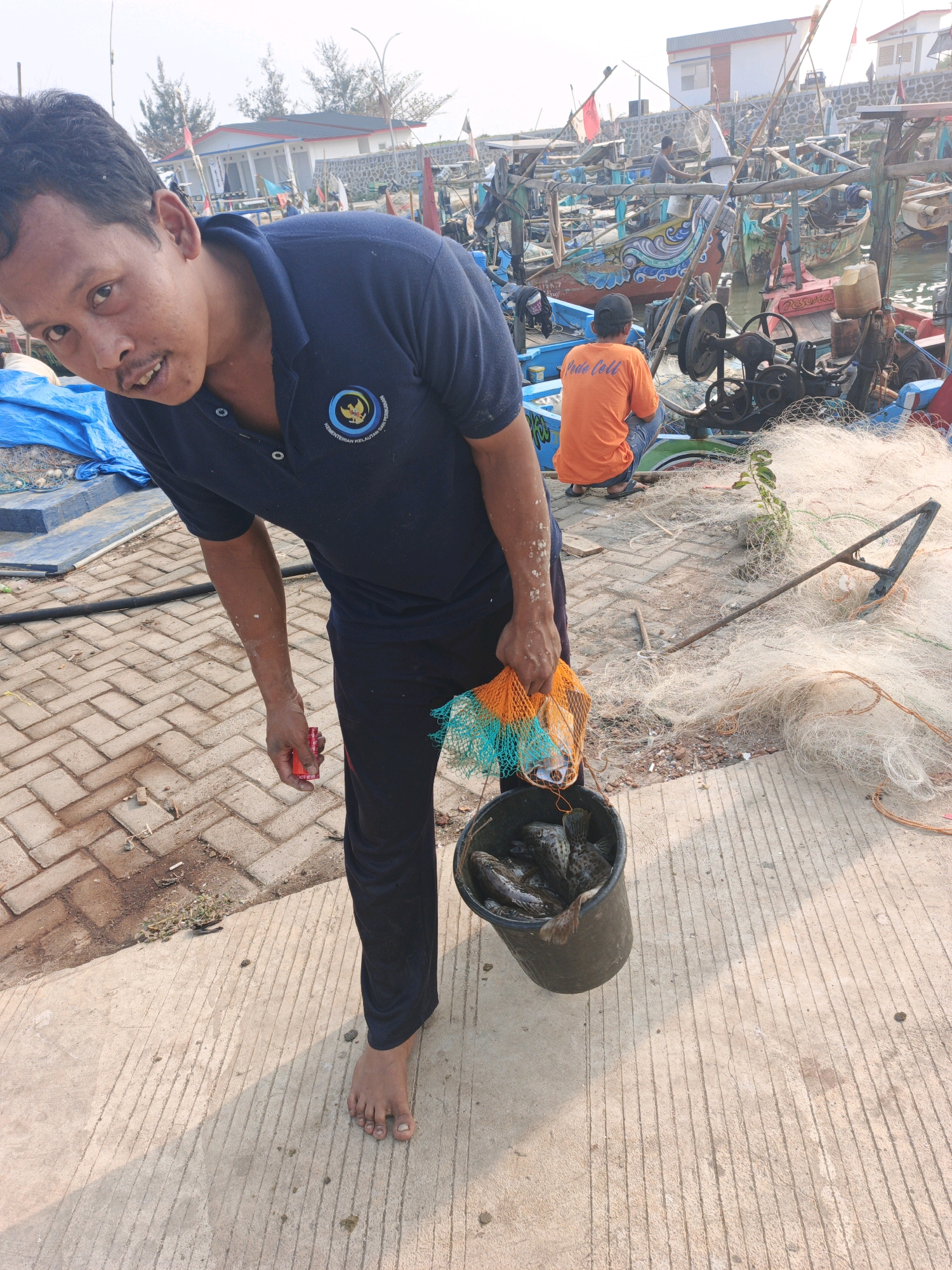 In Unknown, Indonesia, a man with short black hair and a mustache, wearing a dark blue polo shirt with a government emblem and dark pants, stands on a paved walkway near a body of water. He is holding a red object in his left hand and a net bag containing fish in his right. He is looking towards the camera with a neutral expression. A black bucket filled with fish rests at his feet. In the background, several colorful fishing boats are moored, and fishing nets are spread out on the ground. Another person, wearing an orange t-shirt, is visible in the middle ground, crouching near the boats. The sky is bright and appears to be daytime. The environment suggests a fishing port or harbor.
