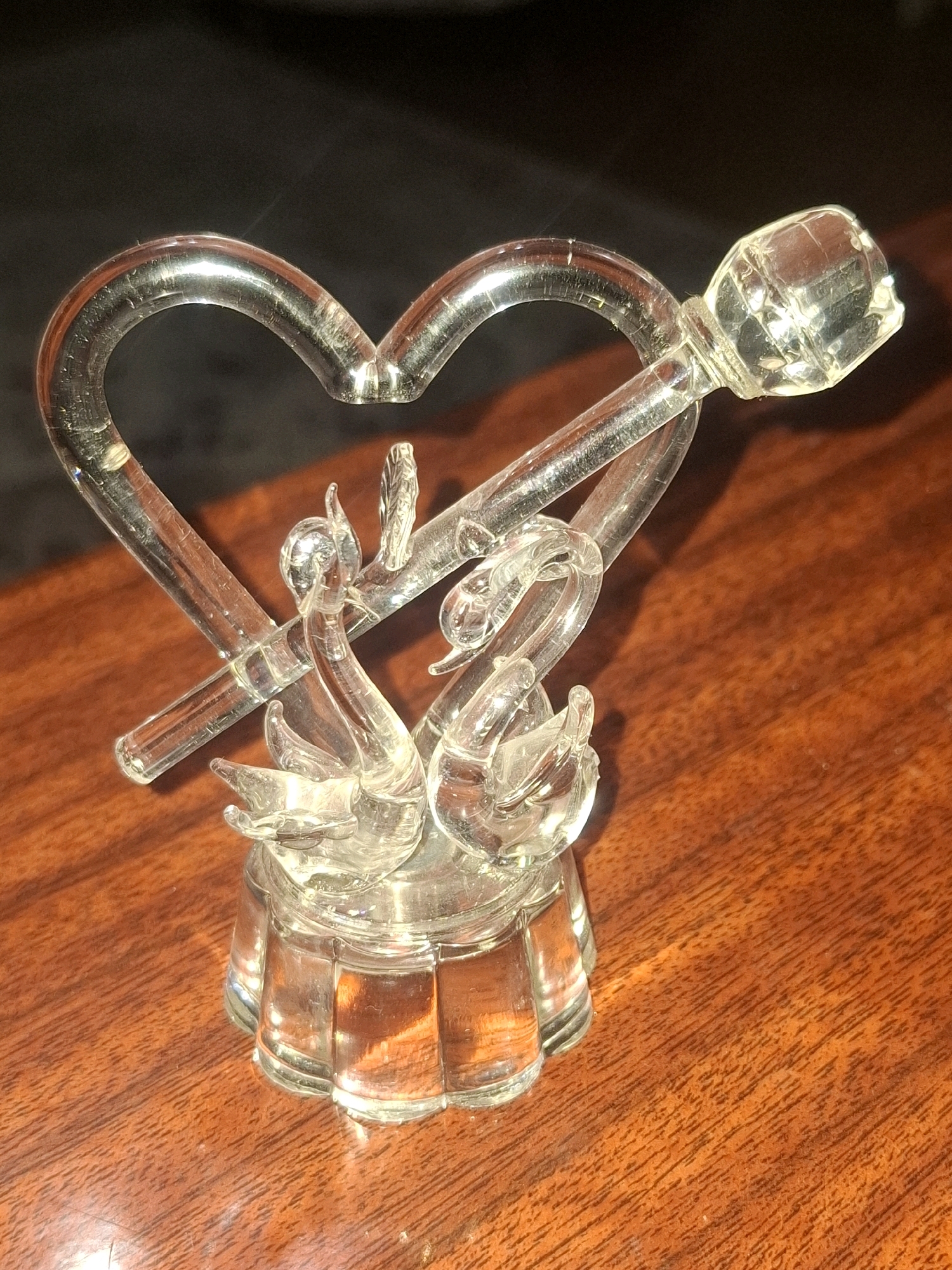 A clear glass figurine depicting two swans facing each other, forming a heart shape, is displayed on a polished wooden surface. A stylized arrow pierces through the heart, with a faceted clear glass embellishment at one end. The figurine rests on a fluted glass base. The background is dark and out of focus. The setting appears to be an indoor domestic environment, such as a table or shelf. There are no people or animals present.