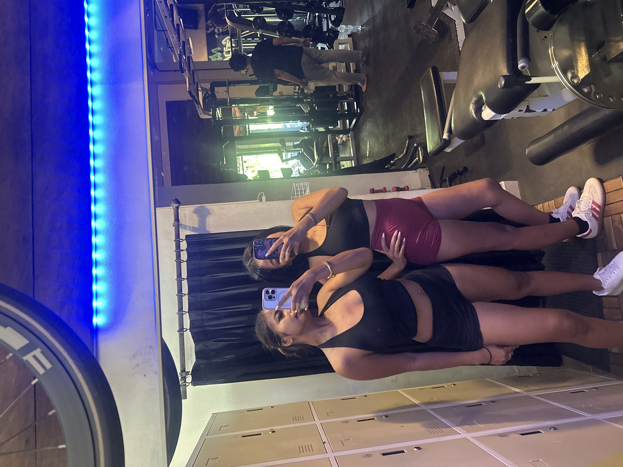 Two young women are taking selfies in a gym locker room, reflected in a mirror. The woman on the left is wearing a black halter-neck crop top and black shorts, holding a phone in her right hand. The woman on the right is wearing a black sports bra and maroon high-waisted shorts, holding a phone in her left hand. Behind them are rows of lockers. The gym itself is visible in the background with workout equipment, including weight machines and benches. A man in a black t-shirt and light-colored pants is seen using a weight machine in the distance. The environment suggests a typical fitness facility, with bright LED lighting above. The overall scene depicts a moment of casual activity and self-expression within a gym setting, possibly in Denpasar, Indonesia, as indicated by the context of the query.
