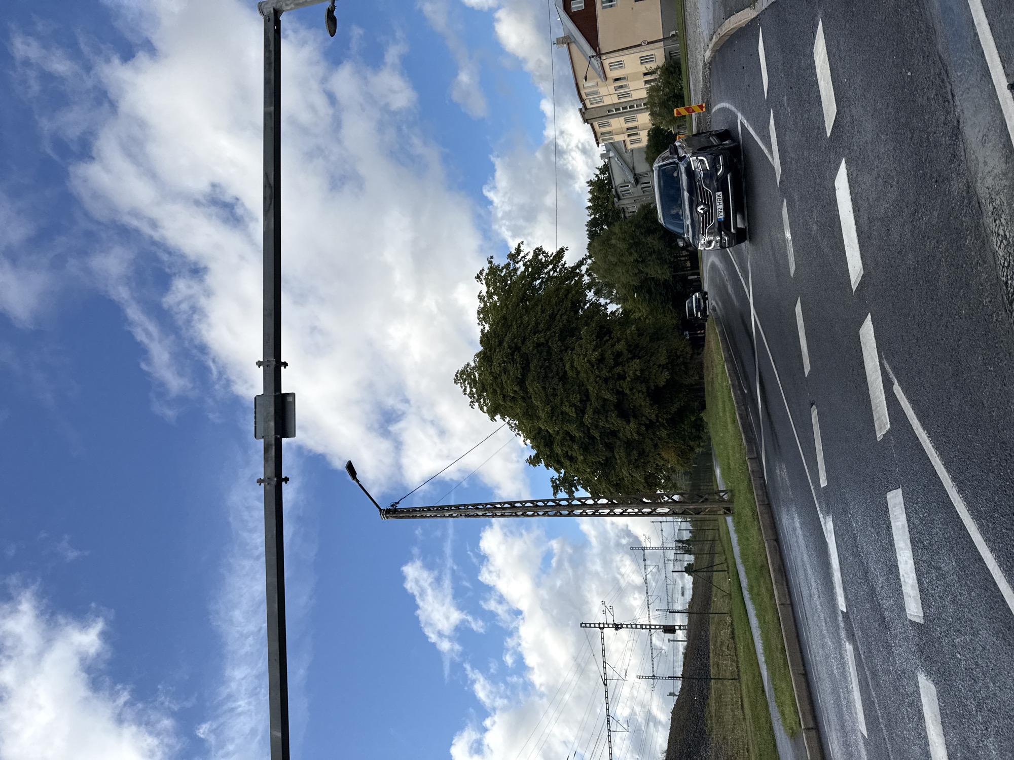 This image captures a street scene in Tallinn, Estonia, on a cloudy day, likely in the late morning or early afternoon. The primary focus is a dark-colored car, possibly a Renault, with license plate "792 HBK", driving on a wet asphalt road marked with white dashed lines. The road curves to the right, bordered by a grassy verge and a gravel embankment. In the background, several buildings with light-colored facades and dark roofs are visible, suggesting a residential or urban area. Mature trees with dense green foliage line the street, casting shadows.
The scene is dominated by a large, dark metal pole on the left side of the frame, which appears to be part of a lighting or electrical infrastructure. Another, more intricate metal structure, likely a power line pylon, is visible further in the background, along with numerous overhead wires. The sky is a mixture of blue and white clouds, with some wispy formations indicating movement. The wetness of the road suggests recent rainfall. The overall impression is of an ordinary day in an urban environment, with the infrastructure of a city and its transportation systems being prominent elements alongside the natural and built environment. No people are clearly visible in the image.