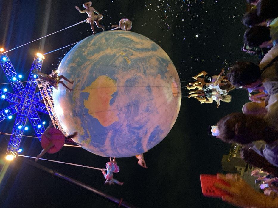 At night, a large globe depicting a world map hangs suspended. Multiple figures in costumes are attached to it, some dangling from ropes, others riding swings projecting from the globe's surface, and one standing on top. To the left, a blue star-shaped structure is illuminated. A crowd of onlookers with phones in hand are on the right, likely capturing the performance. Confetti is in the air adding to the celebratory scene.