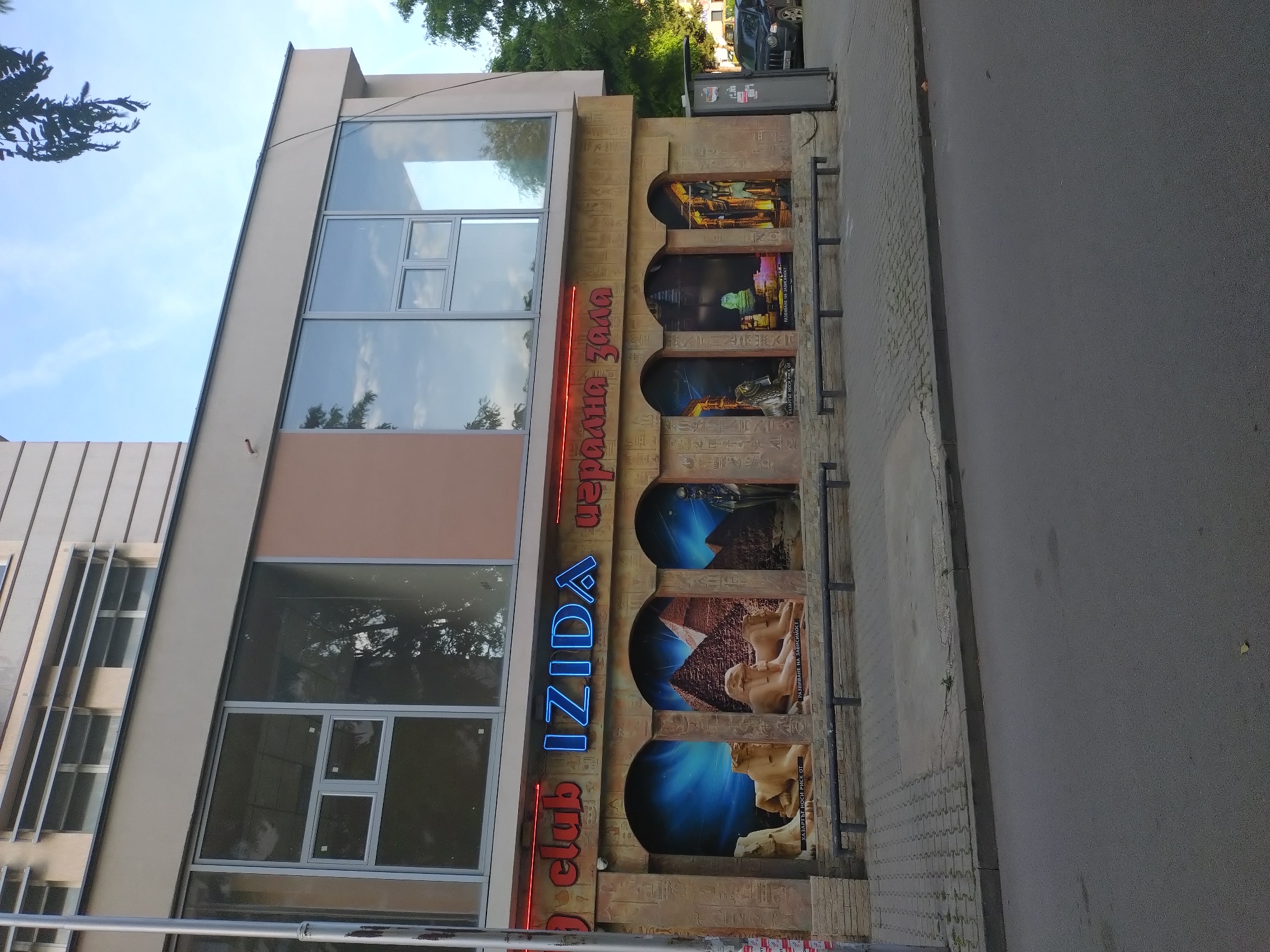 The image depicts the exterior of "IZIDA club & ердания зала" located in Stara Zagora, Bulgaria. The building has a modern facade with large windows and a decorative side panel adorned with Egyptian-themed imagery displayed in a series of arched alcoves. The text "IZIDA club" is displayed vertically in blue and red neon lighting. The side panel features images of Egyptian artifacts and landscapes with hieroglyphic-like carvings. The building fronts onto a gray paved street. A vehicle is visible parked further down the street in the background.