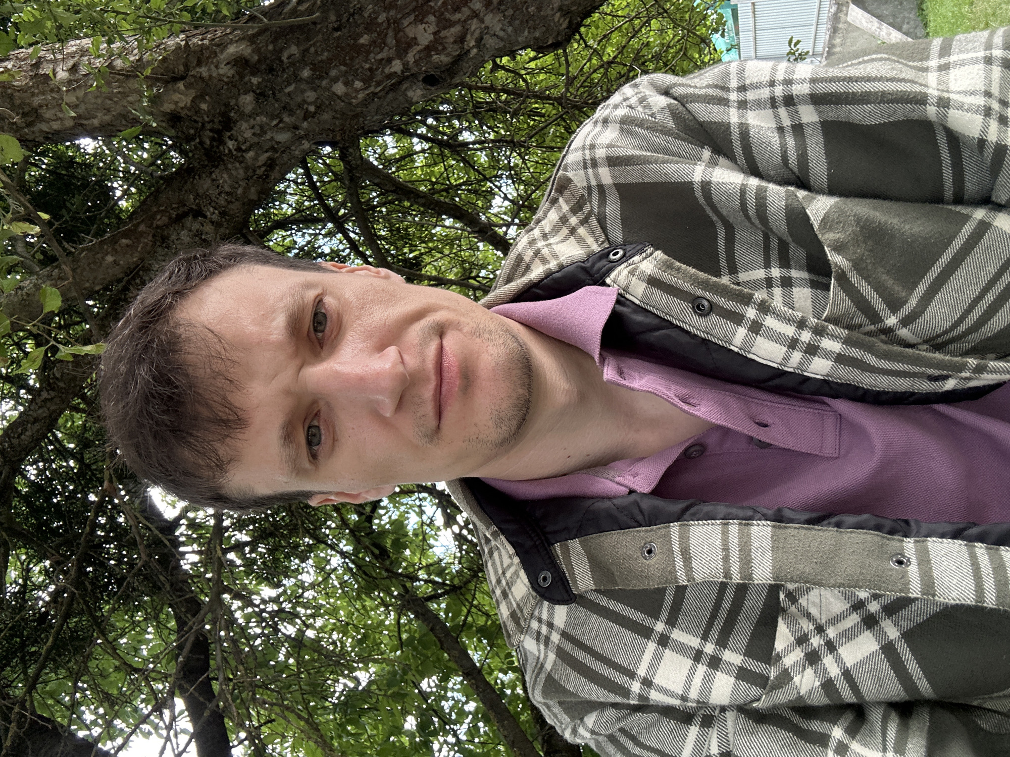 A man is pictured from the chest up, outdoors, with a tree and foliage behind him. He has short, dark brown hair and is wearing a plaid jacket over a pink polo shirt. The man is looking directly at the camera with a neutral expression. The background consists of a large tree trunk and branches with green leaves, suggesting a park or wooded area. A glimpse of a metal fence and a concrete structure can be seen on the right side of the frame, indicating a residential or urban fringe environment. The lighting suggests it is daytime. The location is specified as Makiv, Ukraine.