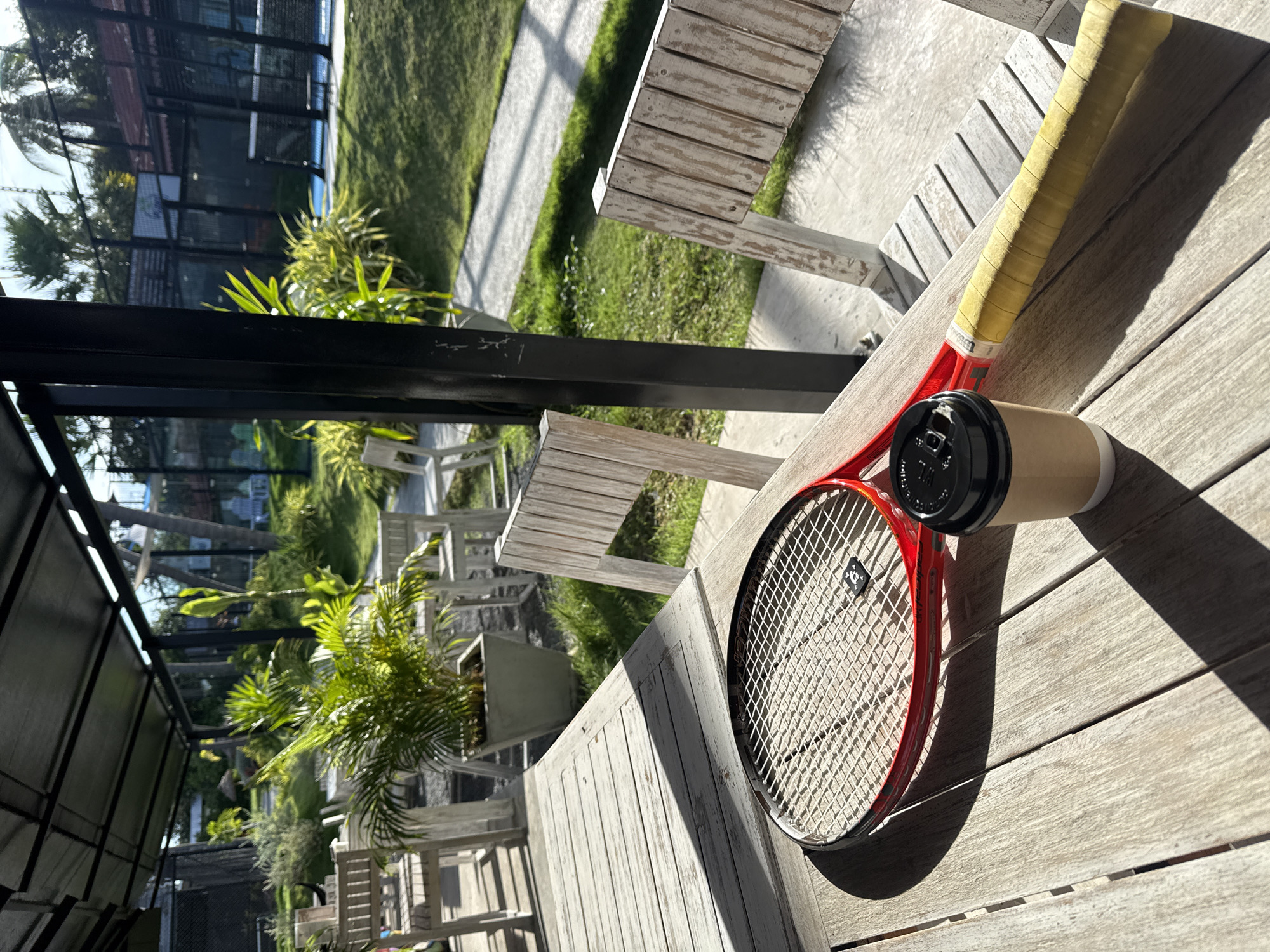 A red tennis racket rests on a light-colored wooden table next to a disposable coffee cup. The racket's strings are visible, and a small black accessory is attached near the center. The handle of the racket is wrapped in yellow tape. In the background, there are white wooden chairs, potted plants, and a glimpse of outdoor tennis courts with fences. The scene appears to be an outdoor seating area in Baan Plai Laem, Thailand, with palm trees and lush greenery suggesting a tropical climate. The lighting indicates it is daytime, with shadows cast across the table and the ground.