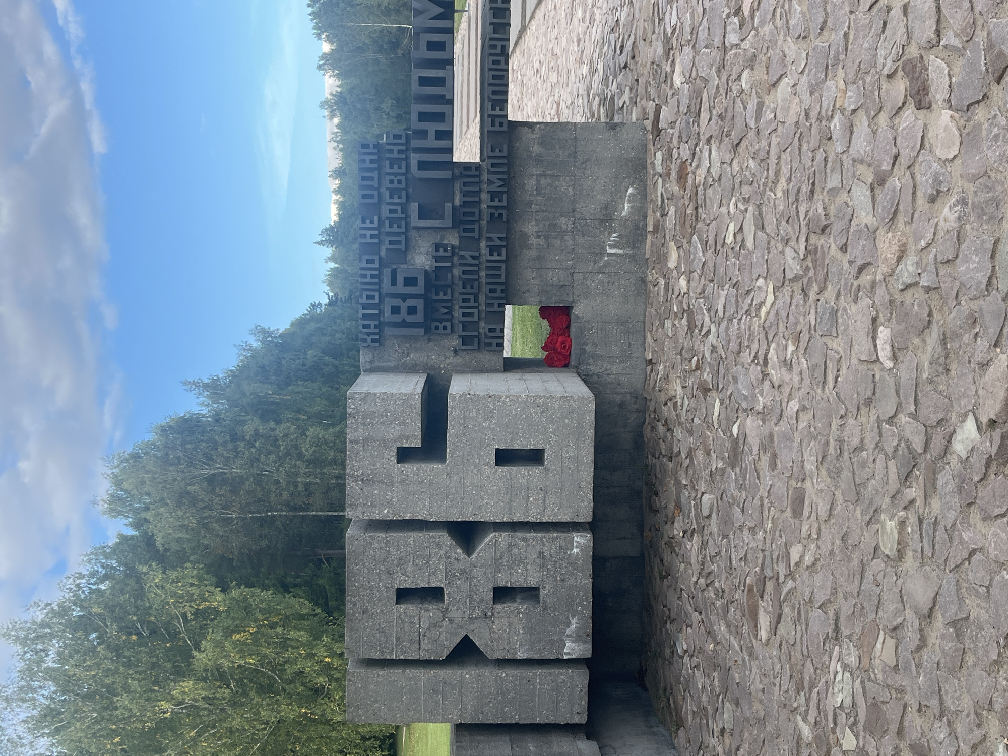 The image shows a monument with large concrete block numbers "186" in the foreground. Behind the numbers, there is a section of the monument with text inscribed in Cyrillic. The text appears to be a memorial inscription. To the left of the "186" monument, there are red flowers placed in a recess of the concrete structure. The ground in front of the monument is paved with irregular, flat stones. In the background, there are trees and a blue sky with some clouds. The scene is outdoors, with a clear day. The monument is located in the city of Каменскі сельскі Савет, Belarus.