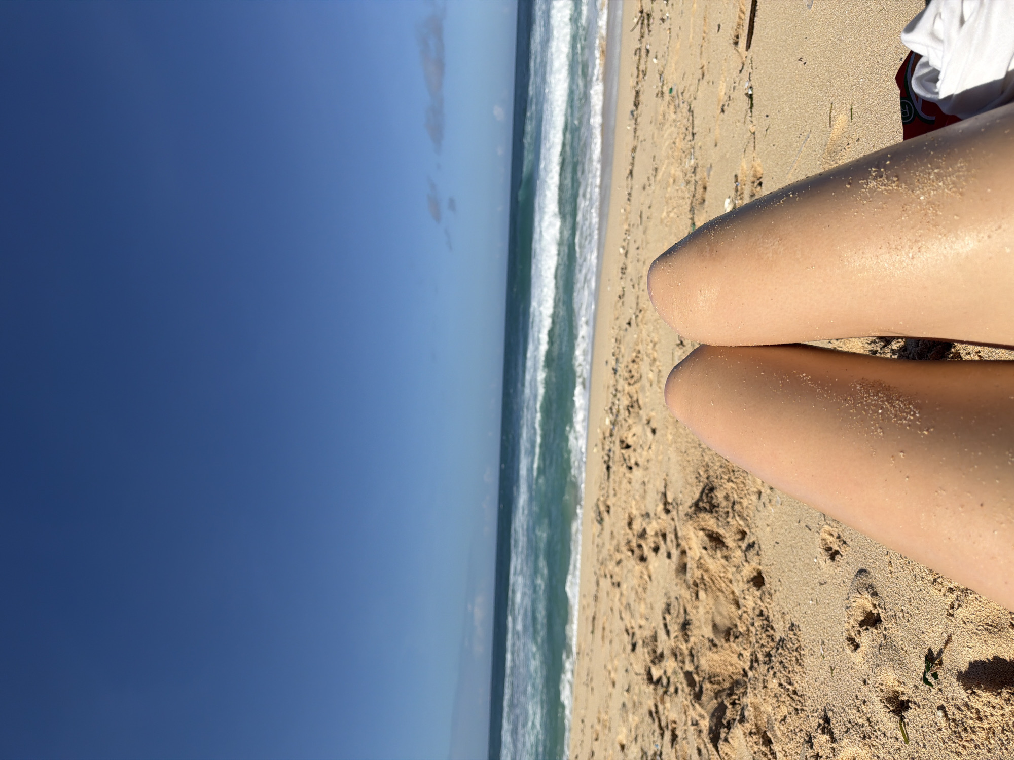 The image shows a view from the perspective of a person lying on a beach in Kuta Selatan, Indonesia. The sky is a clear, deep blue with a few faint clouds. The ocean is a turquoise color, with white waves breaking along the shoreline. In the foreground, the sun-tanned legs of the person are visible, resting on the sand, which appears to be a golden tan color. Some sand is stuck to the legs. Near the legs, sand is disturbed in patches.