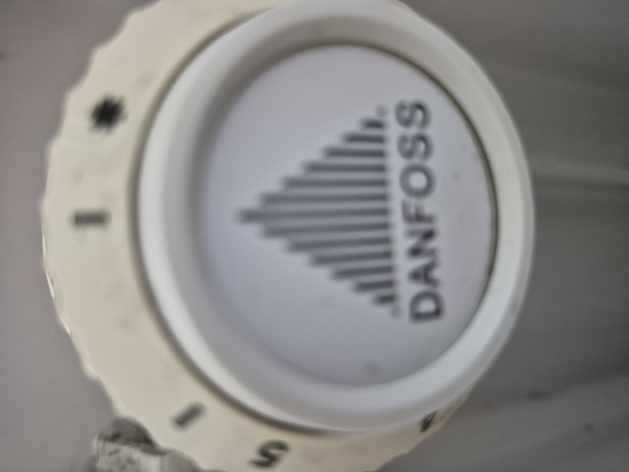 The image shows a close-up of a Danfoss thermostat dial. The dial is cream-colored with black markings. The numbers 1 and 5 are visible on the outer edge of the dial, along with other unreadable markings. The center of the dial features a graphic of vertical bars that decrease in height, resembling a sound wave or temperature indicator, and the word "DANFOSS" is printed below it. The thermostat appears to be mounted on a light gray surface, possibly a wall or appliance. The lighting is diffuse, and the image is slightly out of focus.