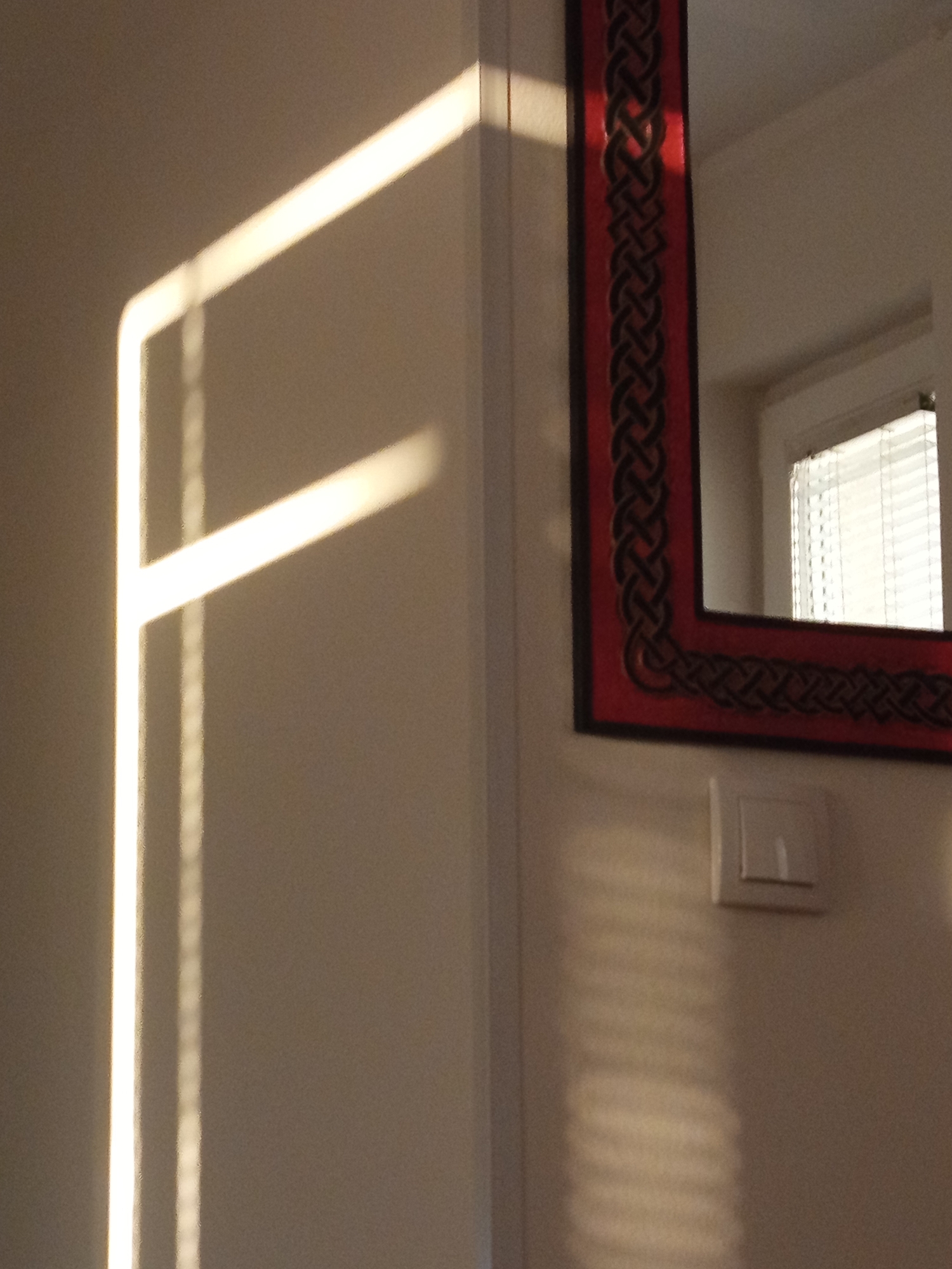 A sunlit, vertical wall is partially illuminated by sunlight streaming through blinds, casting a pattern of horizontal stripes across the surface. To the right of the wall, a decorative mirror with a red frame and an intricate black knot pattern is mounted. The mirror reflects a window with blinds. A light switch is visible on the wall below the mirror. The scene appears to be indoors.