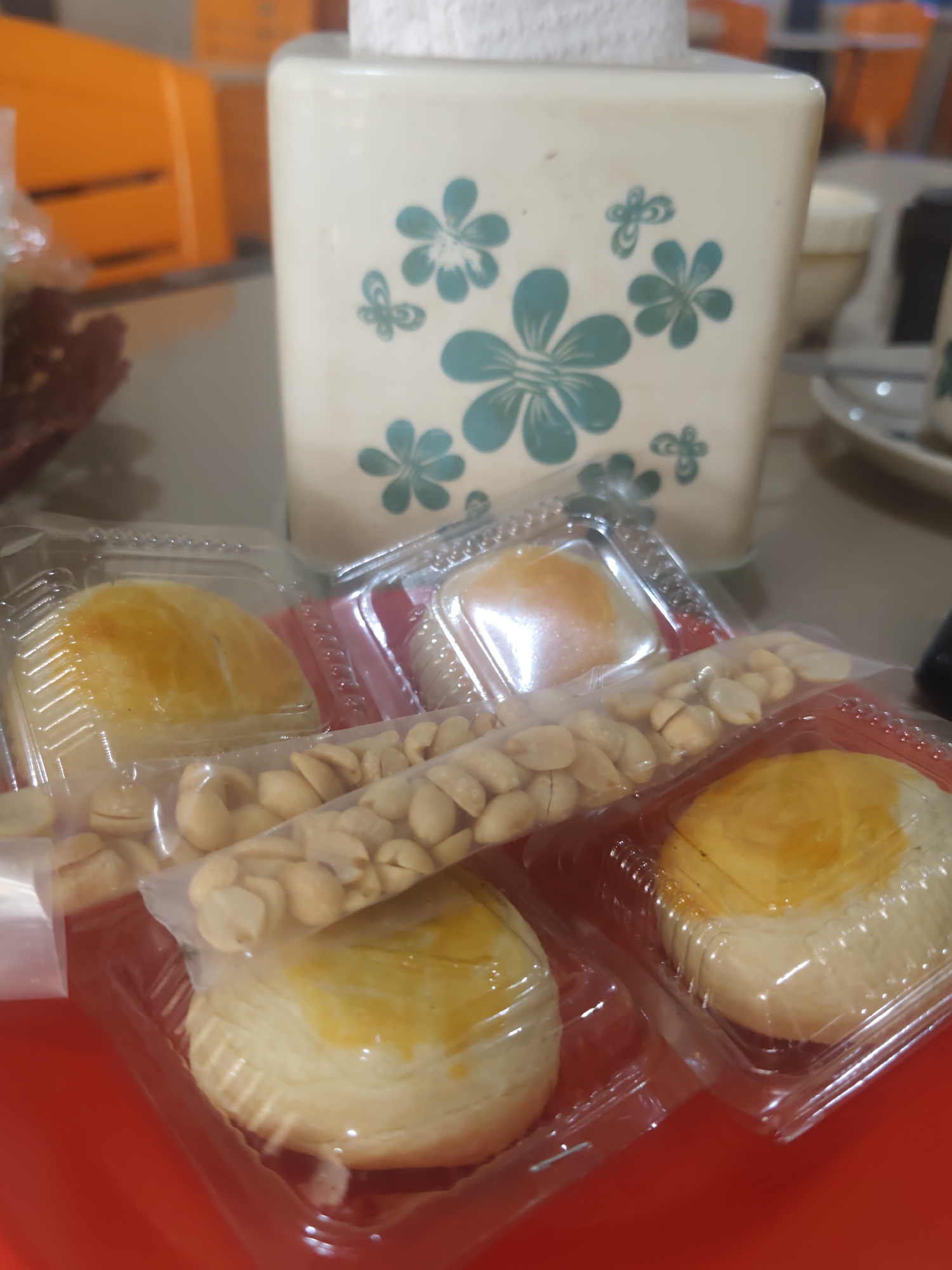 In Pematang Reba, Indonesia, a close-up shot captures a collection of pastries and peanuts arranged on a red tray. The pastries, individually packaged in clear plastic containers, exhibit golden-brown flaky crusts topped with a bright yellow filling. A handful of roasted peanuts, also in a plastic bag, is placed across the pastry containers, hinting at a snack or dessert offering. In the background, a cream-colored tissue holder adorned with light blue floral patterns stands prominently, suggesting a casual dining or home setting. The scene is illuminated by soft lighting, casting a gentle glow on the food items and the surrounding elements.