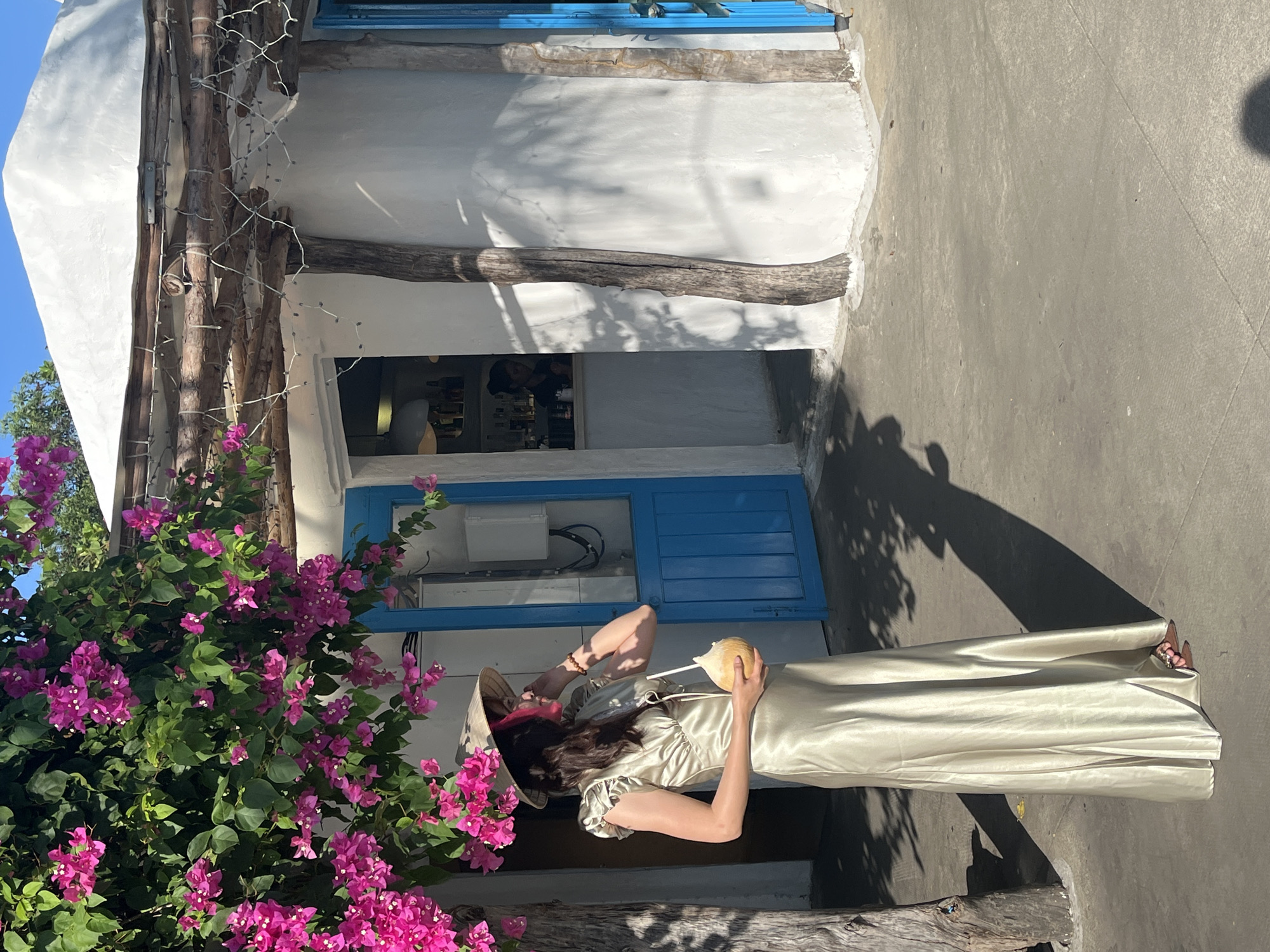 A woman in a conical Asian hat and long dress stands on a gray concrete surface in front of a blue doorway. She is holding a coconut in her right hand. A large bougainvillea plant with magenta flowers is on the left of the frame. The building behind her has white walls and a blue trim around the doorway. Inside the doorway, people are visible. There is a wooden beam above the doorway. The location is not specifically identifiable as being in the city of Đà Nẵng, Vietnam based on visual information.