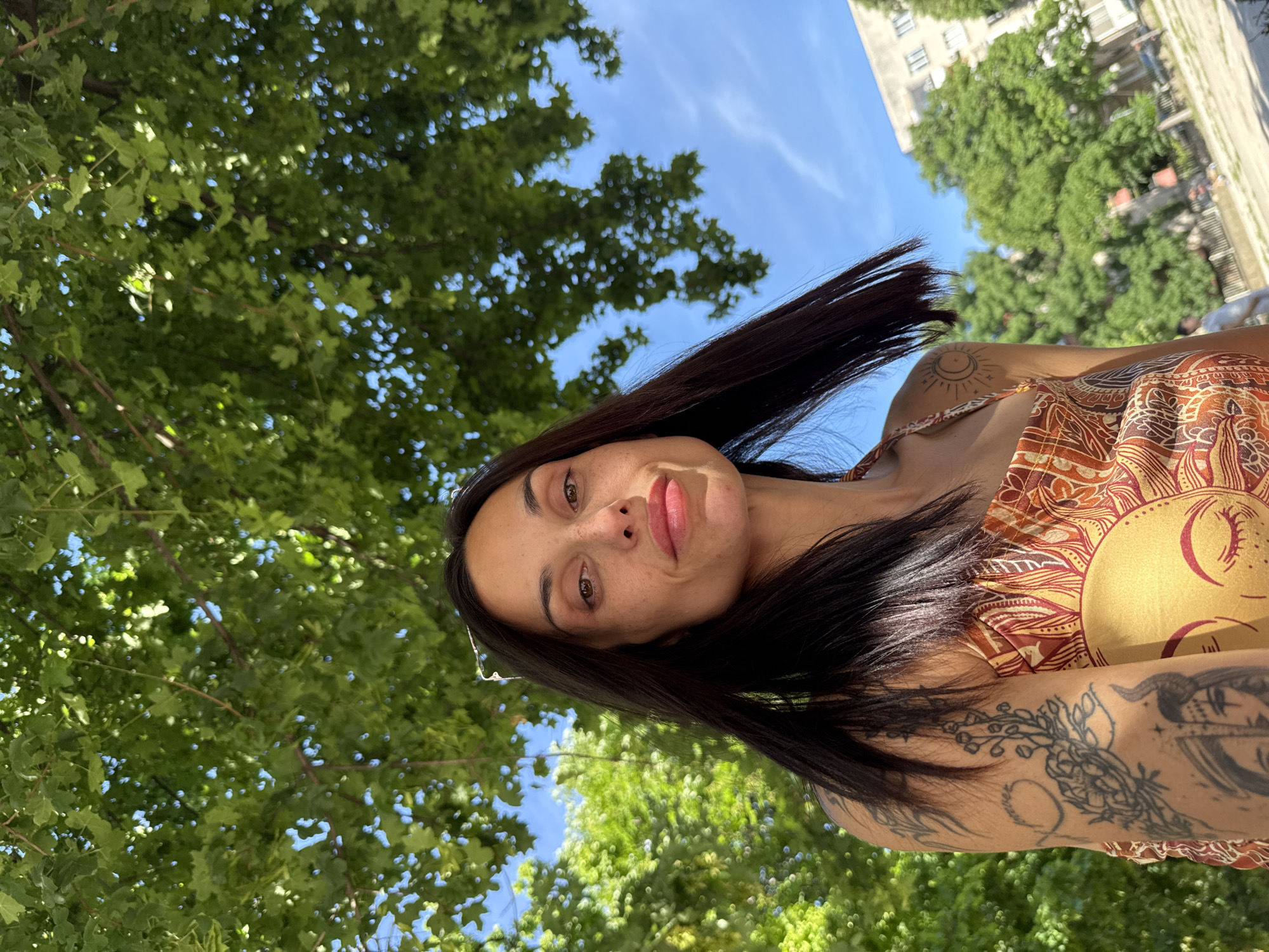 The image features a woman in Pleven, Bulgaria, likely outdoors near green foliage and a building visible in the background. She has dark hair and brown eyes, and is wearing a patterned top or dress, with visible tattoos on her arm and shoulder. The weather appears sunny, as indicated by the clear blue sky and light filtering through the leaves.