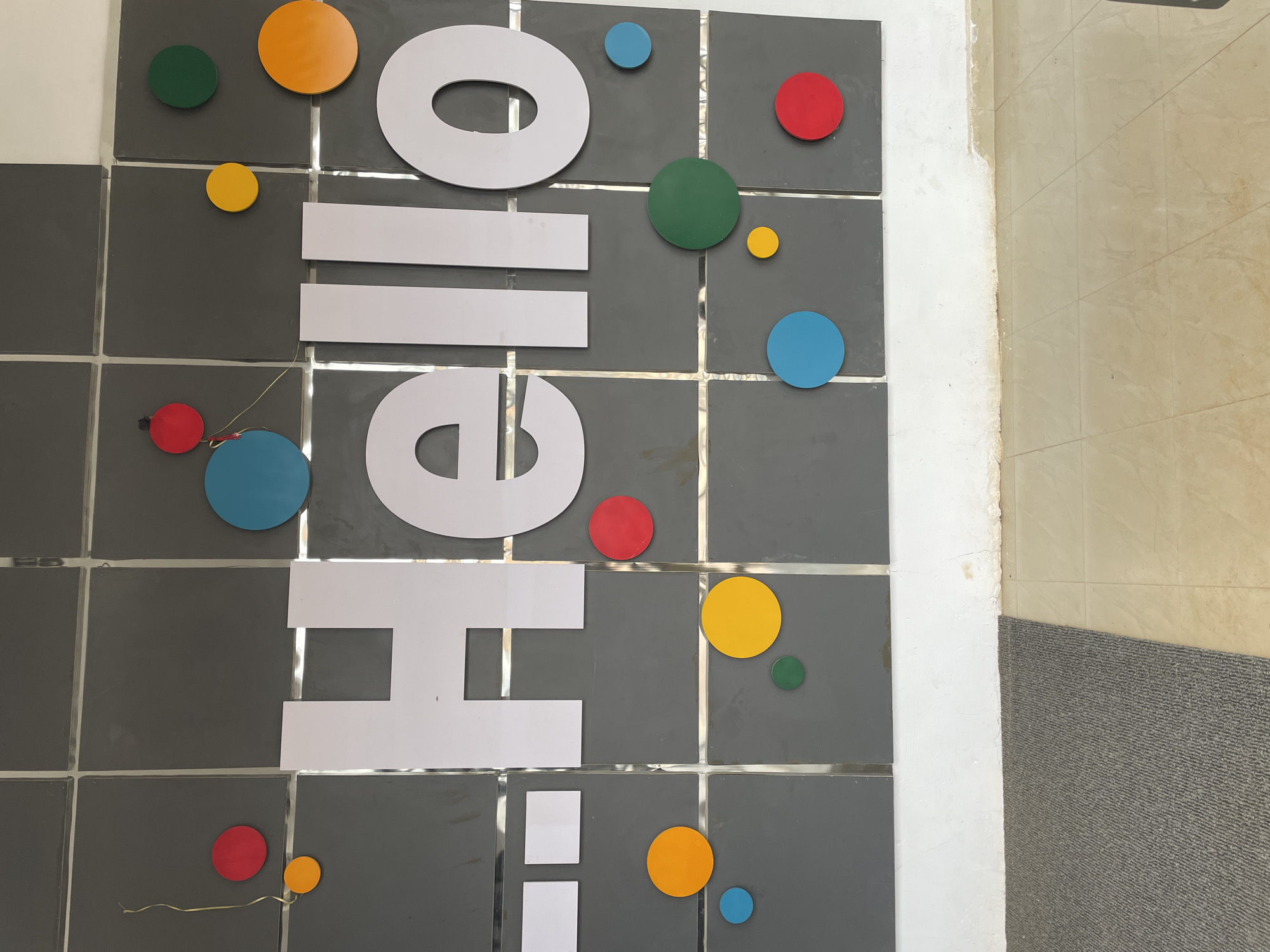 The image displays a decorated wall featuring large, white, vertically stacked letters spelling "HELLO HI" in a bold, sans-serif font. These letters are mounted on a background of dark grey square tiles, which are separated by shiny, reflective silver strips.
Scattered around the white text are numerous circular cut-outs of varying sizes, colored brightly in red, blue, yellow, green, and orange. These circles appear to be decorative elements, possibly resembling balloons or abstract art, contributing to a cheerful aesthetic. A thin, light-colored wire is visible near a red circle in the lower-middle section of the image, suggesting some of these decorations might have an electrical component.
To the right of the tiled wall, there's a section of a plain white wall with a somewhat uneven edge where it meets the tiles. Below this, a light-colored tiled floor is visible, extending towards the bottom right corner of the frame, where a textured, grey doormat or rug is partially shown. The scene depicts an interior space, possibly an entrance or common area in Jos, Nigeria, designed with a welcoming and modern feel. No human activity is depicted.
