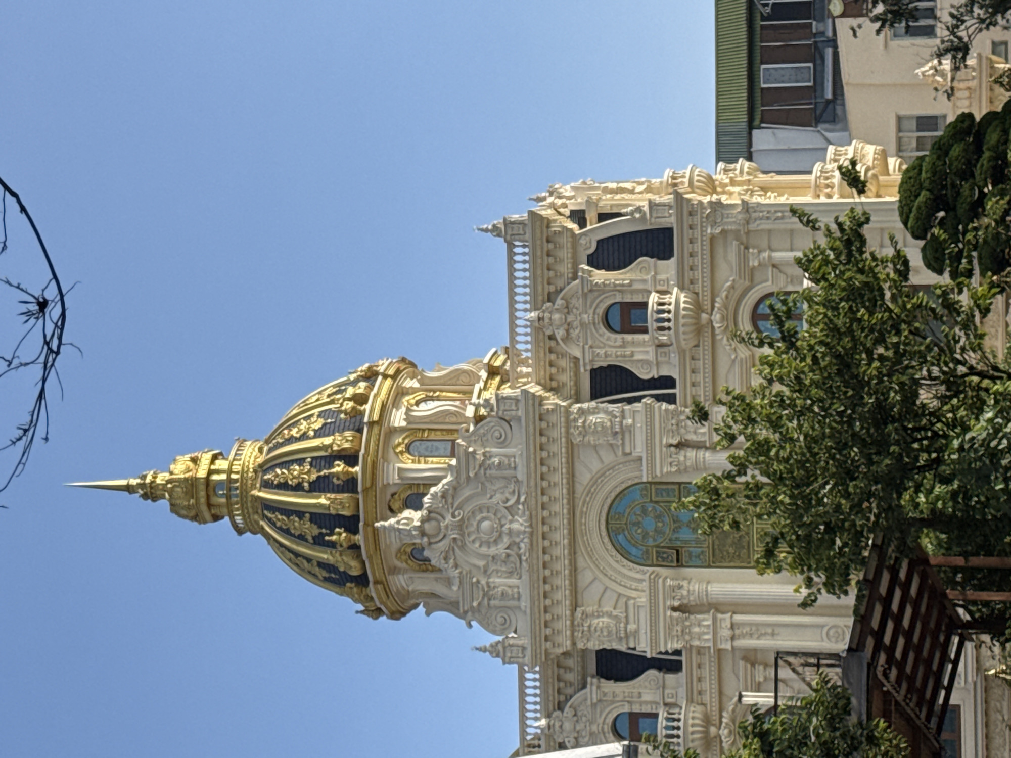 The image shows the exterior of a grand, ornate building with a gold dome and intricate white stonework, set against a clear blue sky. The building features classical architectural elements such as columns, arched windows, and decorative carvings. In the foreground, lush green trees partially obscure the lower part of the building. The architectural style and dome suggest a building of historical or cultural significance. This building is Dinh Bao Dai, also known as the Summer Palace of Emperor Bao Dai, located in Da Lat, Vietnam. The palace was built in 1933 and served as the summer residence for Emperor Bao Dai, the last emperor of Vietnam. It is a prime example of French colonial architecture, with a blend of European styles and Vietnamese influences. The palace is situated on a hill overlooking the city of Da Lat, and its grounds are well-maintained with manicured gardens and lush vegetation, as depicted in the image.