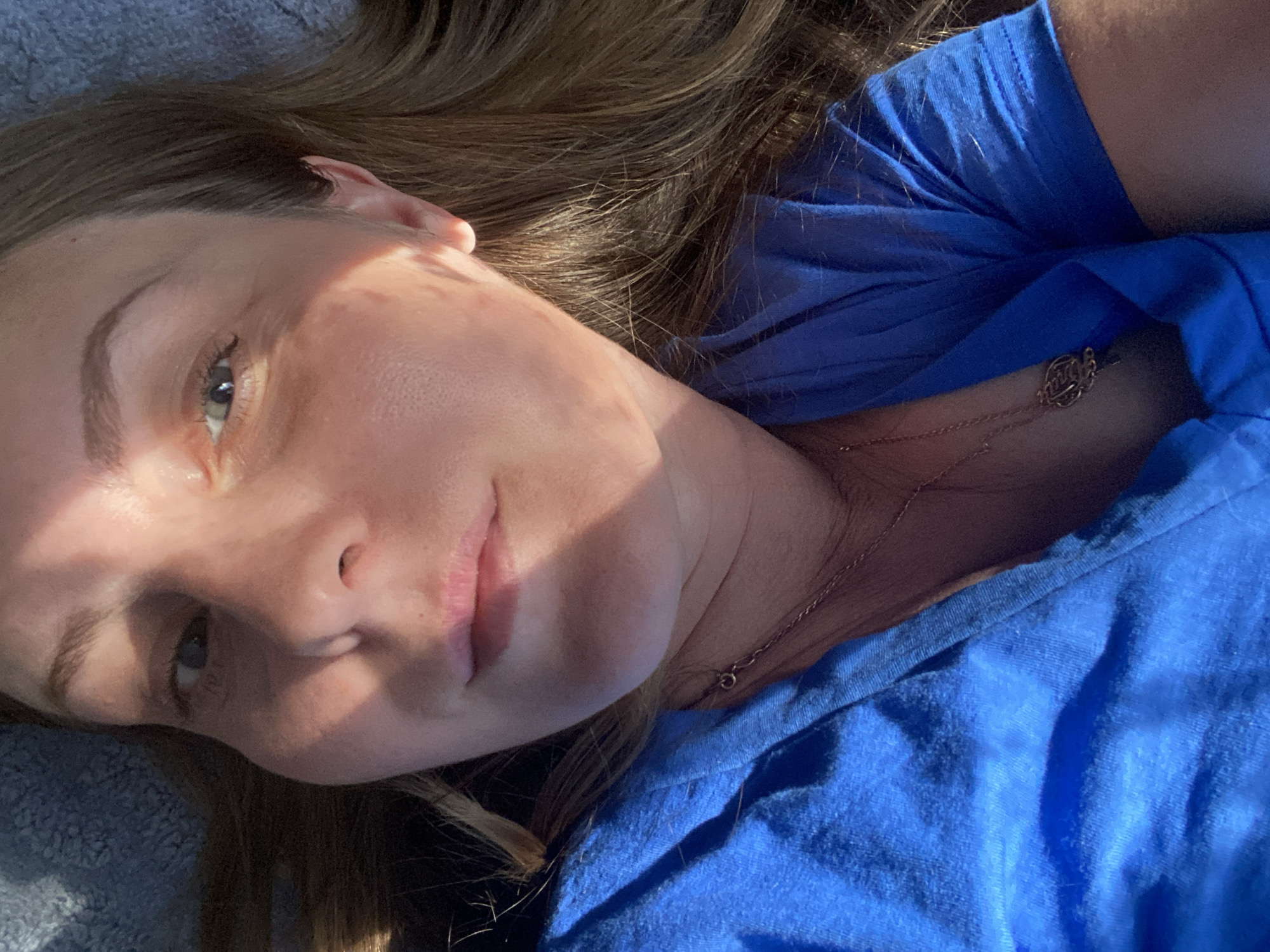 A selfie taken from a high angle shows a woman with long, brown hair and fair skin. She is wearing a blue t-shirt and a delicate necklace with a circular pendant. The lighting suggests it is daytime, with sunlight casting a shadow across her face, highlighting her features, including her eye, nose, and lips. The background appears to be a soft, textured surface, possibly a blanket or carpet, in a muted blue-grey color. There is no indication of the city of Noviy, Russia, or any specific environmental context beyond the immediate surroundings captured in the frame.