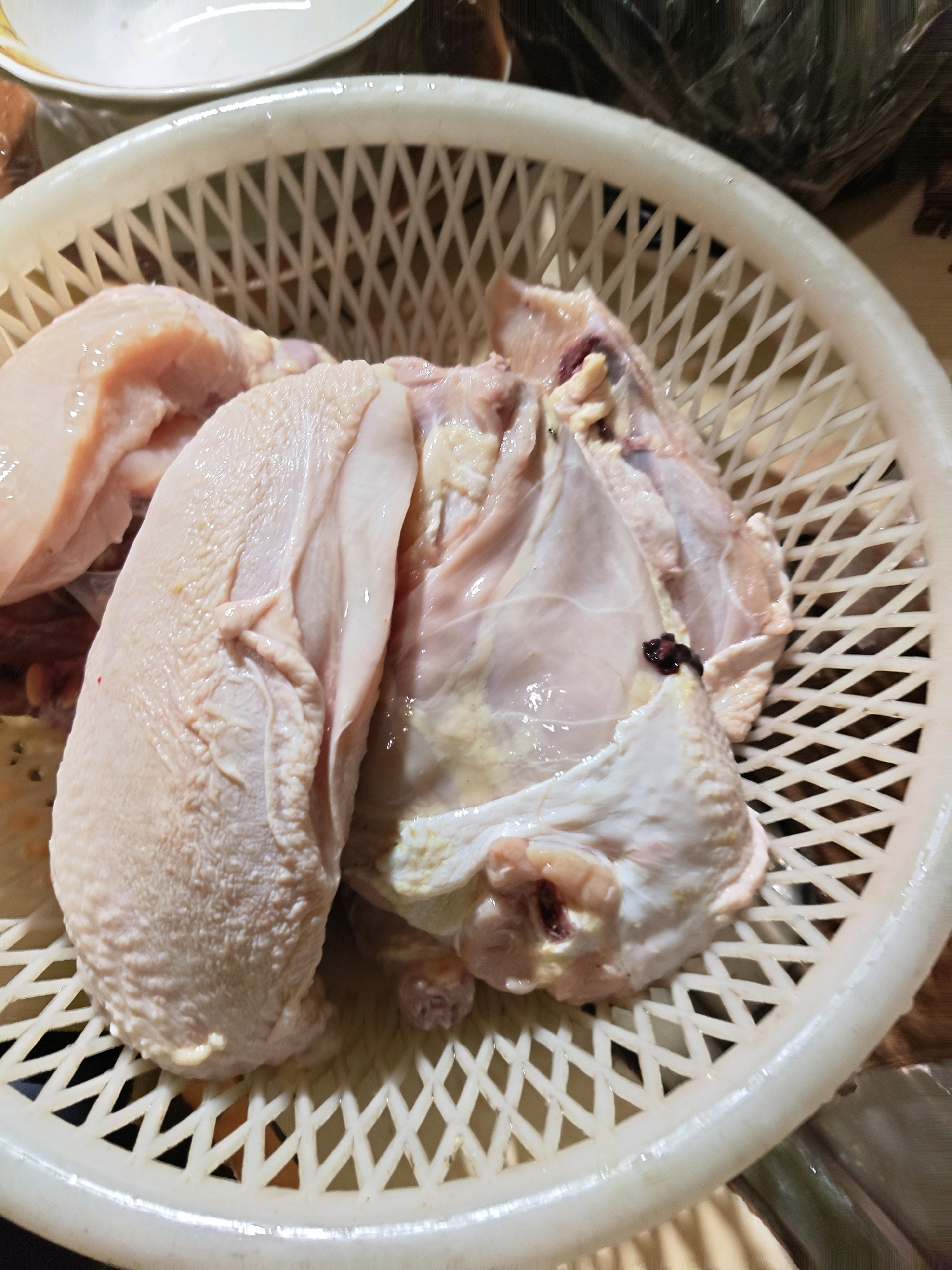 A close-up, top-down shot shows raw chicken pieces in a white plastic basket. The chicken is light pink and pale yellow, with some darker red and black marks visible. The basket has a criss-cross pattern and is sitting on a surface that appears to be a wooden table. In the background, a white plate and some dark green leafy vegetables in plastic wrap are partially visible. The lighting is somewhat dim, creating shadows within the basket. The image appears to be taken indoors, possibly in a kitchen in Sumberwono, Indonesia.