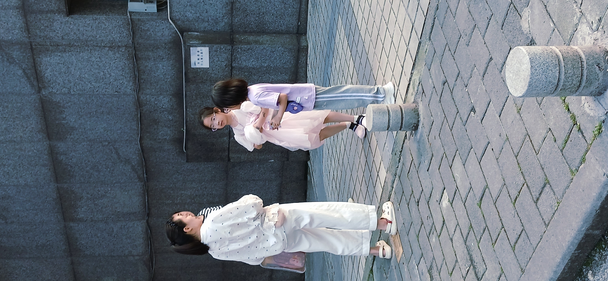 The image depicts three individuals: an adult female and two children (likely girls). The woman is wearing a white, patterned top, white pants, and white sandals. One child wears a pink dress and the other a purple shirt and gray pants. The children interact near the woman. They stand on a brick-paved sidewalk adjacent to a gray wall. The scene is presumably in a city environment, potentially in the Jiulongpo District, China. Cylindrical stone barriers line the edge of the sidewalk.