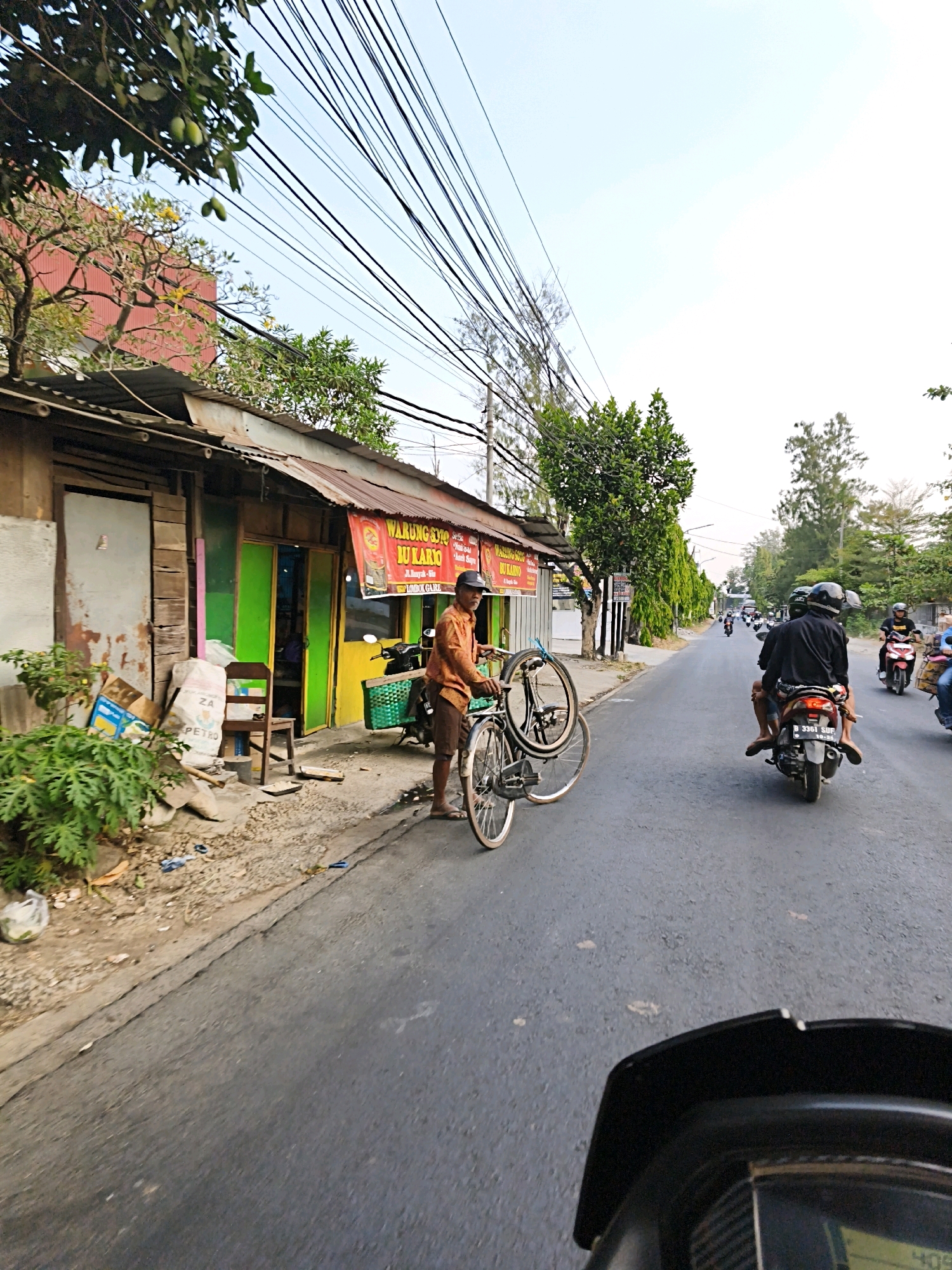 In the city of Ngargorejo, Indonesia, a street scene unfolds with a man standing next to a bicycle on the right side of the road. He is wearing an orange shirt and brown shorts, and a black cap. The bicycle is propped up and appears to be loaded, with its front wheel slightly detached. Behind him, a small shop with a corrugated metal roof and colorful green and yellow doors lines the street. A sign above the shop reads "WARUNG SOPO JU KARYO". The shop's exterior is weathered, with exposed wooden beams and peeling paint. Piles of boxes and bags are stacked near the shop's entrance, and a green plastic basket is attached to a motorcycle parked in front of the shop. The road is paved and shows signs of wear. Several motorcycles are seen traveling down the road in the distance. The sky above is clear and bright.