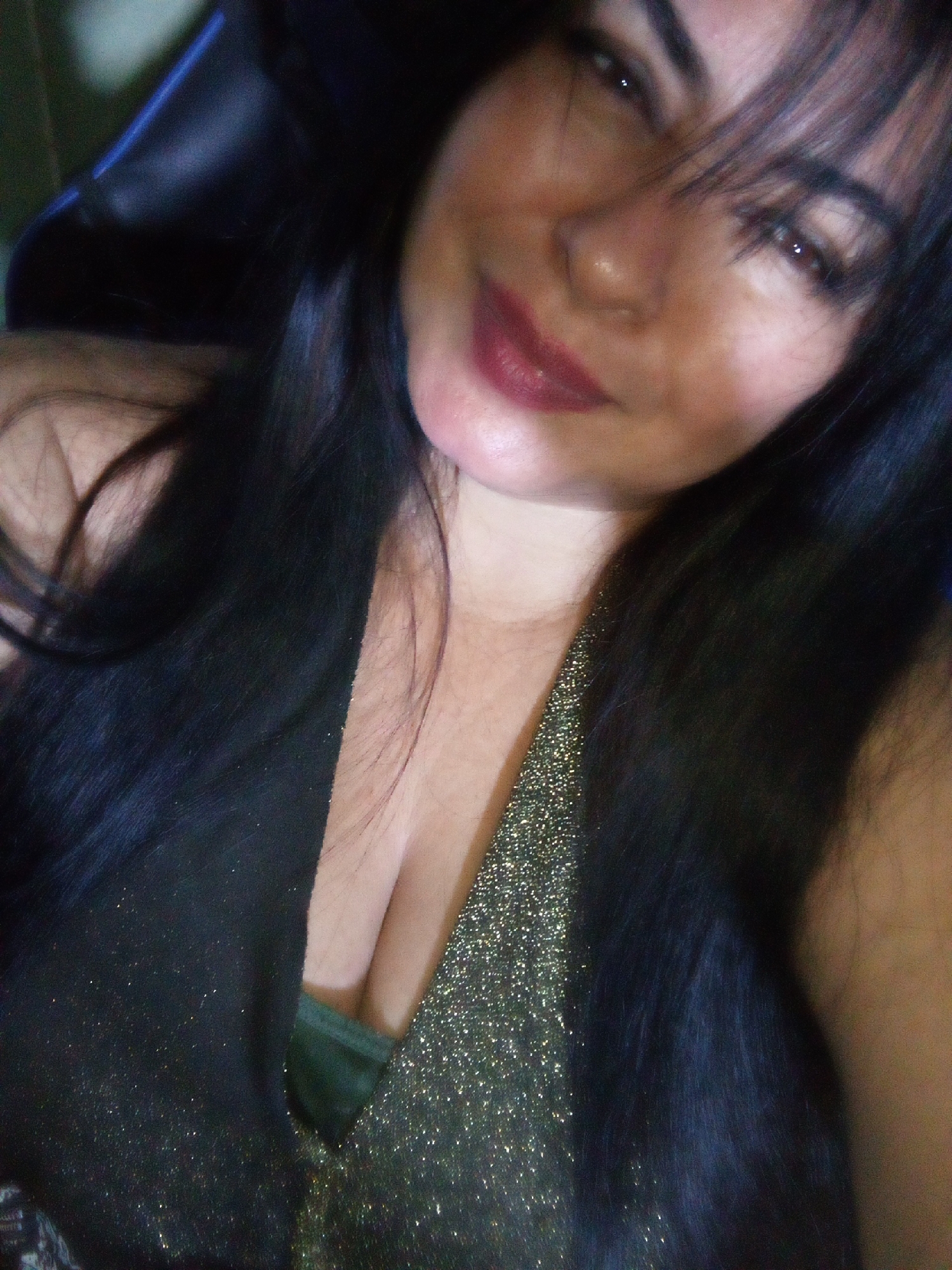 A close-up shot shows a woman with dark hair, gazing at the camera. She's wearing a low-cut top, dark on the left and shimmering gold on the right, suggesting a V-neck design. The background appears to be a dark blue, possibly part of a chair. There are no visible clues to indicate the location in Juan Griego, Venezuela.