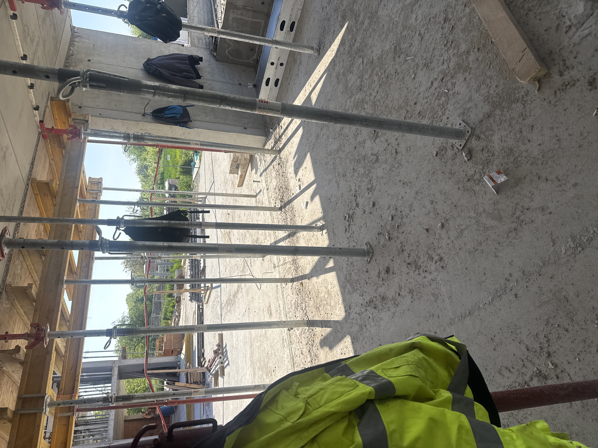 The image depicts an indoor construction site, potentially within a building in Plovdiv, Bulgaria. Concrete walls are visible, supported by numerous vertical metal poles. Several personal items like bags and a high-visibility vest are draped over or near the poles. The floor is rough concrete. Construction materials like wooden planks are scattered around. The daylight suggests the site is either open or has significant openings to the outside.