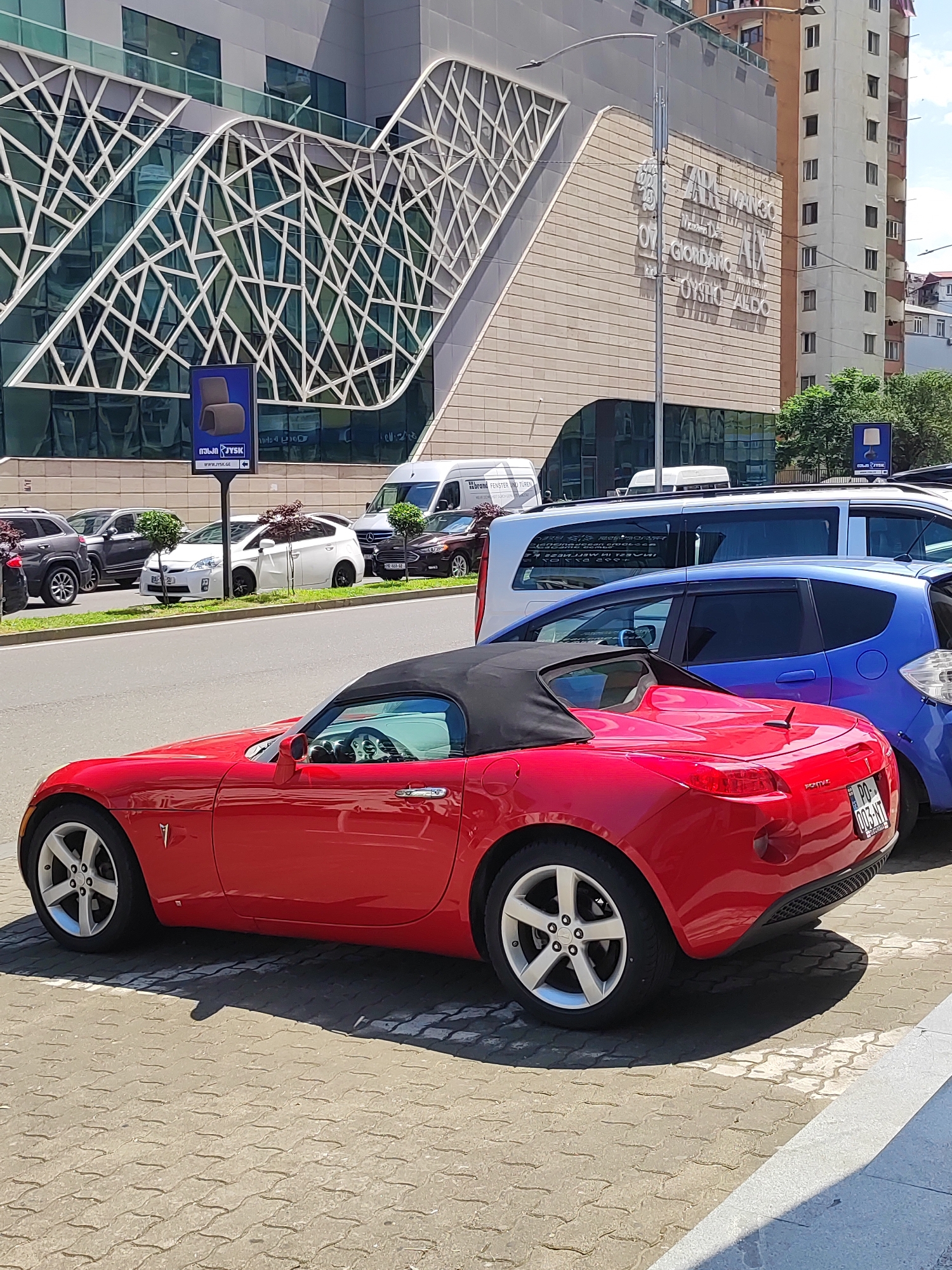 The image shows a red Pontiac Solstice convertible parked on a cobblestone pavement in front of a modern building with a distinctive geometric facade. The building appears to be a commercial establishment, with several brand names visible on its facade, including "MANGO", "GIORDANO", and "OYSHO-ALDO". In the background, other vehicles are parked on the street, including a white Toyota Prius and a blue hatchback. The overall scene is a street view in Batumi, Georgia, likely on a sunny day.