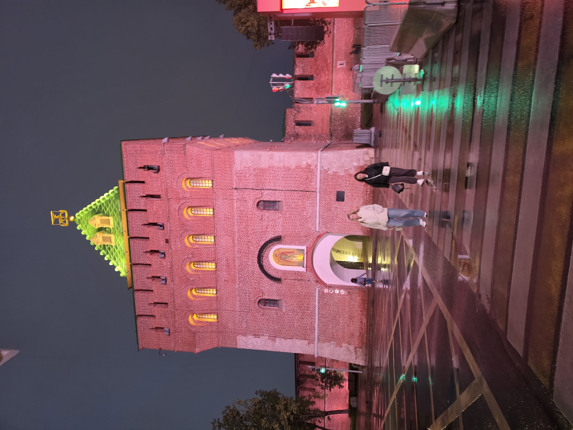 This image captures the Dmitrovskaya Tower of the Nizhny Novgorod Kremlin at night. The tower, constructed of red brick, is illuminated by a reddish-pink light, highlighting its arched windows and ornate green roof. A golden emblem is perched atop the roof. In the foreground, two women walk across a pedestrian crossing, their reflections visible on the wet asphalt. The street is marked with yellow lines forming a grid pattern, suggesting a crosswalk. Traffic lights are visible, with green lights illuminated. The sky is a dark, overcast blue, typical of a night scene. Trees and parts of the Kremlin wall are visible to the left and right of the tower. The overall atmosphere is one of a historical landmark illuminated for nighttime viewing, with a sense of urban activity despite the rain.