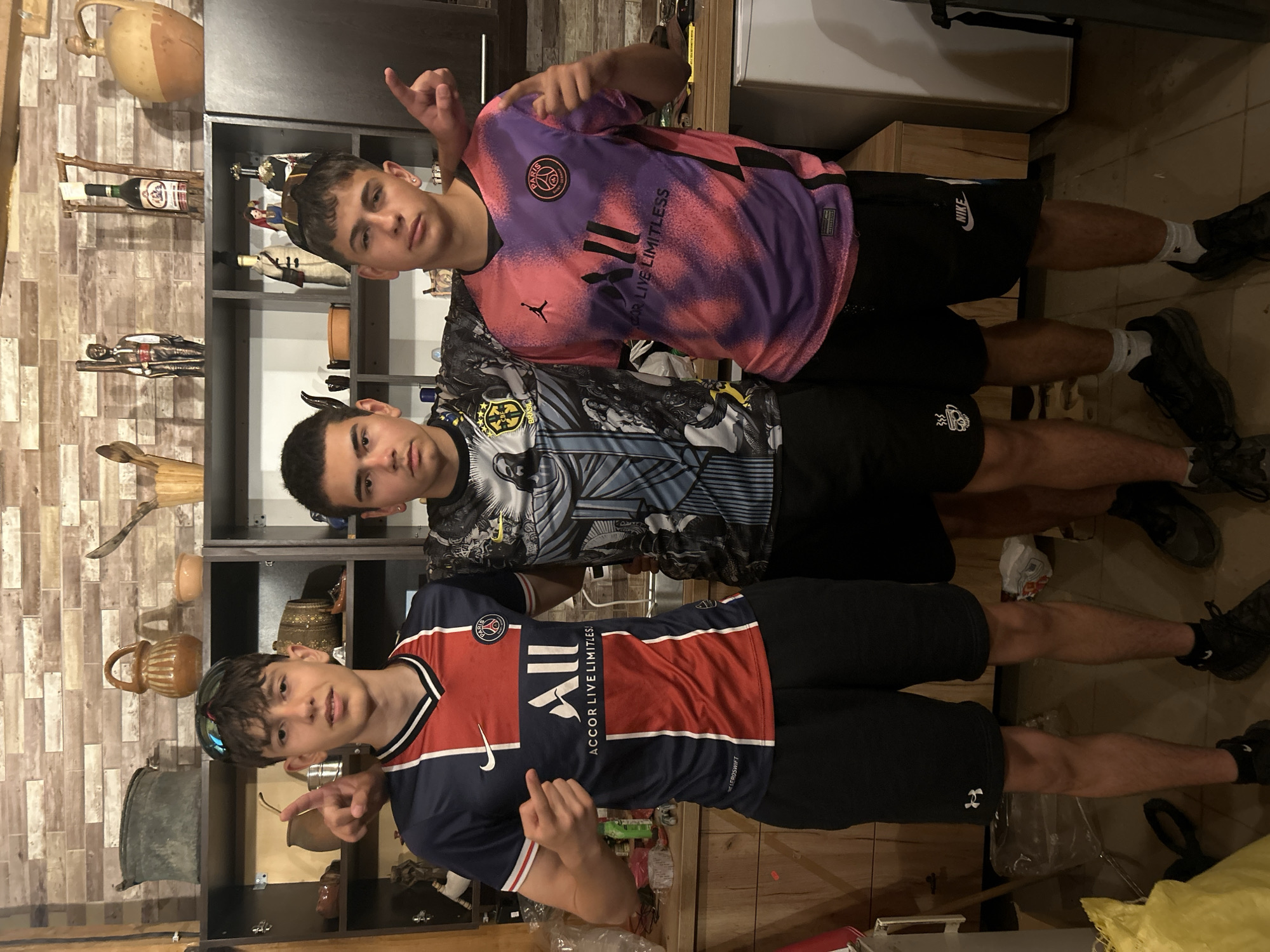 Three young males are posing for a photo indoors, likely in a home kitchen or entertainment area. The male on the left is wearing a blue and red PSG soccer jersey and black shorts, with sunglasses on his head. He is pointing his left index finger upwards. The male in the center is wearing a black and grey soccer jersey with a graphic design and black shorts. The male on the right is wearing a pink and purple tie-dye PSG soccer jersey and black shorts, and is also pointing his right index finger upwards. They are standing in front of shelves filled with decorative items, including vases, bottles, and figurines. The background shows wooden shelving and a tiled wall. The lighting appears to be artificial.