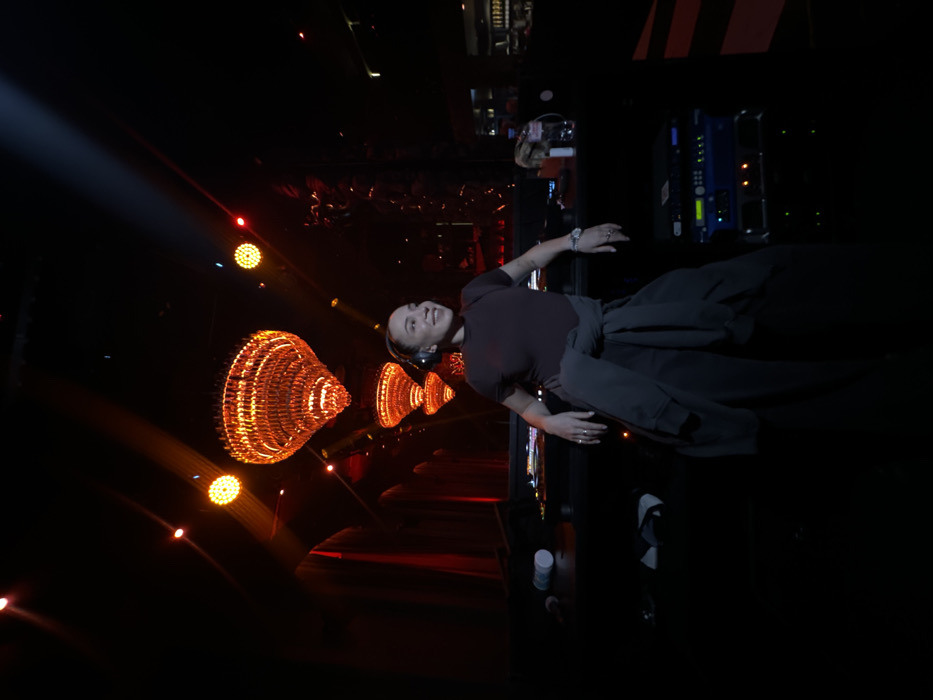 The image captures a female individual, likely a DJ, positioned behind a console in a dimly lit nightclub. The location is identified as Gipsy Club in Moscow, Russia. The woman, appearing to be in her 20s or 30s, has light hair pulled back and is wearing over-ear headphones. She is dressed in a dark, short-sleeved top and dark trousers, with a dark jacket or sweatshirt tied around her waist. Rings are visible on multiple fingers of her left hand, which also bears a wristwatch. She is smiling and looking towards the upper left quadrant of the frame.
The environment is characterized by dark walls and a dark ceiling from which multiple large, ornate chandeliers, composed of numerous small, reflective elements, hang. These chandeliers are illuminated with a prominent orange-red light, creating a warm glow. At least three such chandeliers are visible, receding in a line into the background. Bright, focused spotlights emit white beams from above, cutting through the ambient darkness. Smaller, circular red lights are also visible on the ceiling.
In front of the woman, a dark console or DJ booth is present. On the console surface to her left (viewer's right), a white cylindrical container and other dark objects are visible. To her right (viewer's left), a clear plastic bottle, possibly containing water, and additional items are placed. Below the console, a stack of rack-mounted electronic equipment with illuminated blue and green indicators is visible, suggesting audio or lighting control apparatus. The overall scene is one of a live performance or event setting within a sophisticated, mood-lit venue.