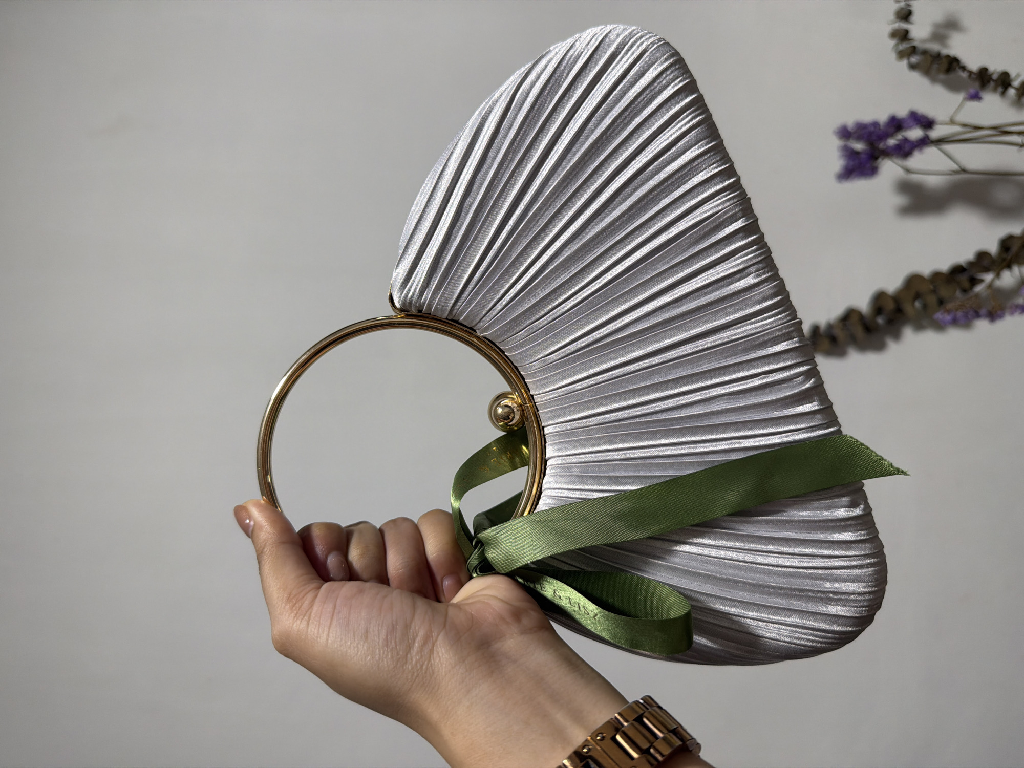 A woman's hand is holding a silver pleated clutch purse with a gold circular handle and green ribbon accents. The purse has a curved, fan-like shape. The woman is wearing a gold watch on her left wrist. In the background, a blurred arrangement of dried flowers with purple accents is visible. The scene is lit with artificial light, creating shadows on the plain, light-colored wall behind the subject. The location is not specified in the image.