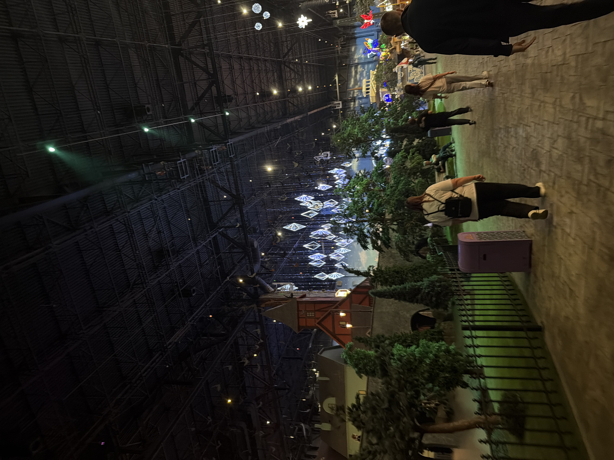 The image shows an indoor scene, likely a shopping mall or entertainment venue, decorated for a festive occasion, possibly Christmas. The ceiling is high and industrial, with exposed metal beams and corrugated panels, suggesting a large indoor space. Strings of lights, some white and sparkling, others forming diamond shapes, are suspended from the ceiling, illuminating the space.
In the foreground, several people are walking along a paved walkway. One person in the very front is seen from behind, wearing a white t-shirt and black pants, carrying a black bag. Other individuals are further down the walkway, also walking away from the camera. The walkway is bordered by artificial trees and decorative building facades, creating a whimsical, almost European-themed environment. A purple trash receptacle is visible to the right.
The overall lighting suggests it is evening or nighttime, as the interior lights are prominent and the exterior light is not visible. The atmosphere appears relaxed and casual, with people strolling through the decorated area. The decorative elements, including the lights and the faux storefronts, create a festive and inviting ambiance. There is no visible text in the image.