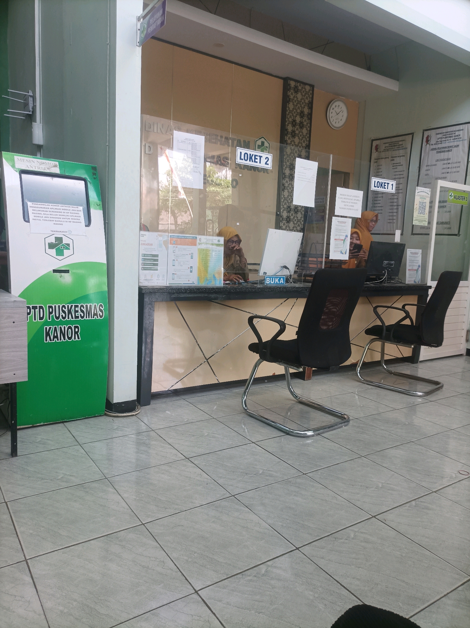 The image depicts the interior of a public health center (Puskesmas) in Tambahrejo, Indonesia. A green kiosk labeled "MESIN NOMOR ANTRIAN" (Queue Number Machine) stands to the left of the frame. Directly behind it, a counter with a glass partition is manned by two female staff members wearing headscarves. One staff member is interacting with a computer, while the other is looking at her phone. In front of the counter, two black mesh chairs with chrome legs are placed on a tiled floor. Various informational posters and signage are visible on the wall behind the counter, including signs indicating "LOKET 1" (Counter 1) and "LOKET 2" (Counter 2). A wall clock is mounted above the counter. The environment appears to be a waiting area or service point within the health center.