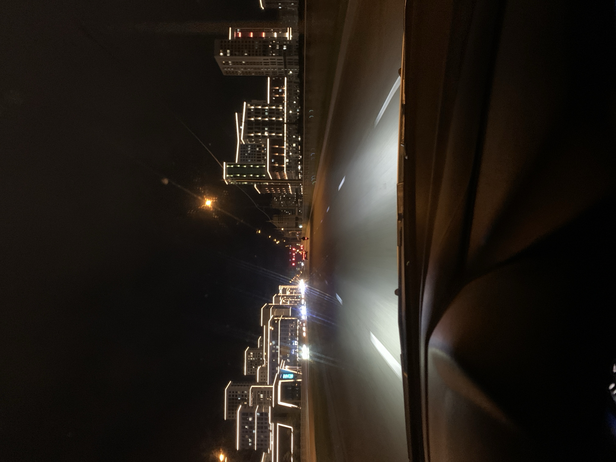 The image depicts a nighttime scene from the perspective of someone driving a vehicle on a multi-lane road. The road is illuminated by the vehicle's headlights, which create a strong beam of light in the center of the frame. The road surface is asphalt, with white lane markings visible.
On either side of the road, there are modern, illuminated buildings. These buildings are multi-story and feature prominent architectural lighting, outlining their edges with a bright, warm-toned light. The buildings appear to be apartment complexes or commercial structures, with multiple windows visible.
In the distance, other vehicles can be seen on the road, with their headlights and taillights creating streaks of light due to the motion blur. The overall atmosphere is that of a developed urban area at night, with significant artificial lighting from both the road and the surrounding architecture. The sky is dark, indicating it is well past sunset.
The specific location is the city of Stavropol, Russia, on a main thoroughfare, likely a highway or a major avenue designed for high-speed traffic. The architecture suggests a relatively new or recently developed part of the city. The perspective is low, typical of a car's dashboard view, with the windshield partially visible at the bottom.