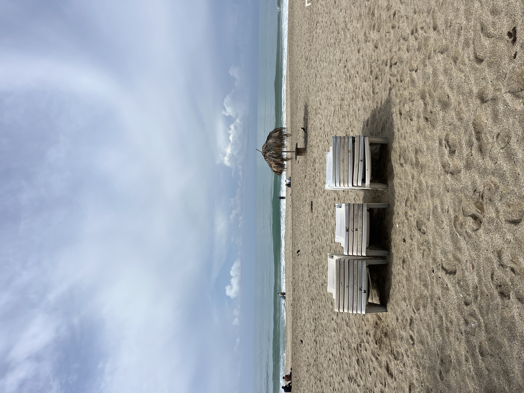 The scene is a beach, oriented vertically in the image, with the sky and sea on the left and the sand on the right. The sky is overcast with clouds, and the sea is calm with small waves breaking along the shore. On the sand, there are stacks of white beach chairs and a thatched beach umbrella. There are a few people visible near the water's edge. The location is Alanya, Turkey.