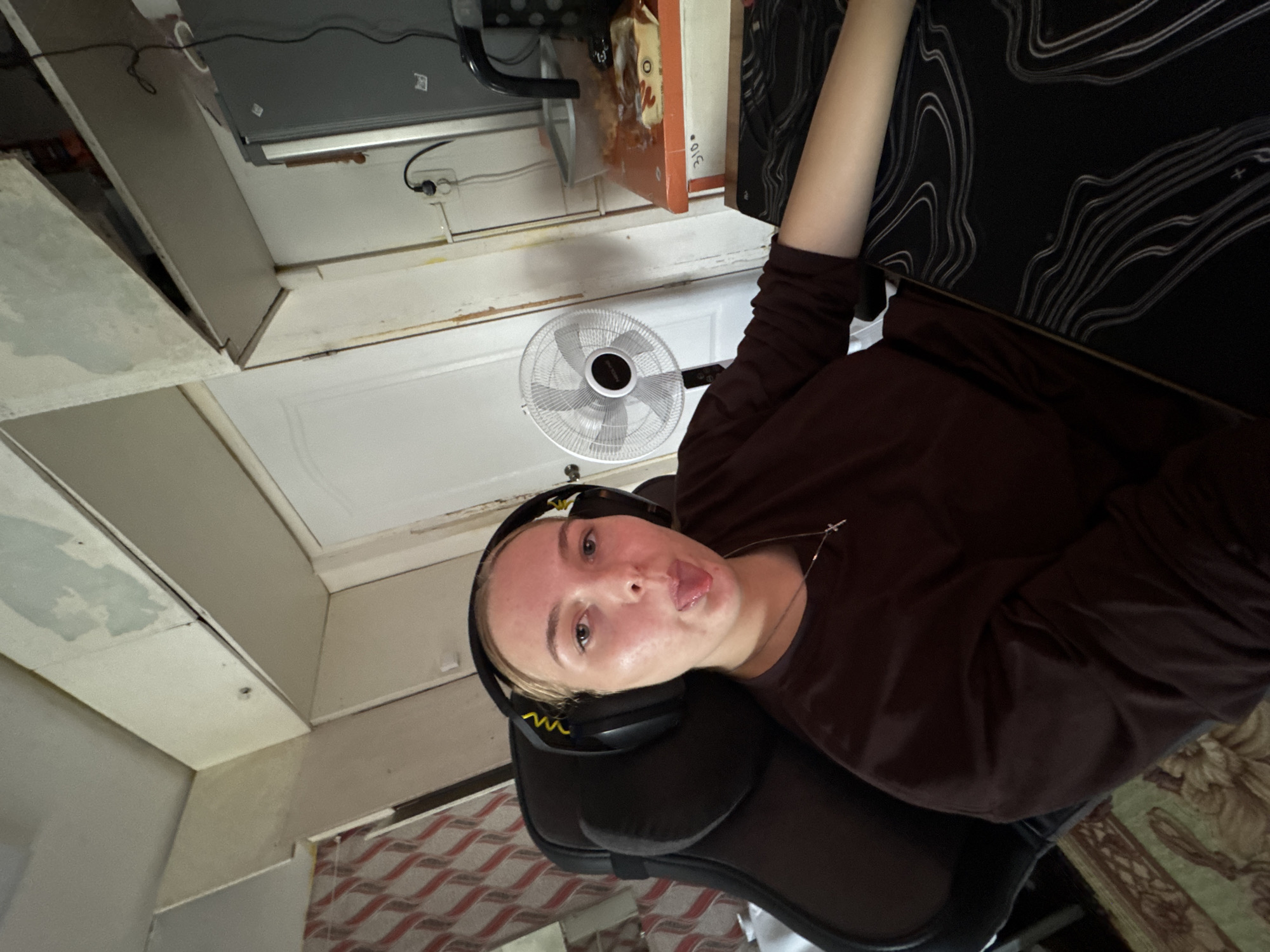 A woman with blond hair is seated in a black gaming chair, wearing a dark brown long-sleeved shirt and black headphones. She is sticking her tongue out towards the camera. A ceiling fan is visible in the background. Above the fan is a door-like structure. The background also shows elements of a cluttered interior with what appears to be damaged ceiling paint. The location is indicated as Perm, Russia, based on external information.