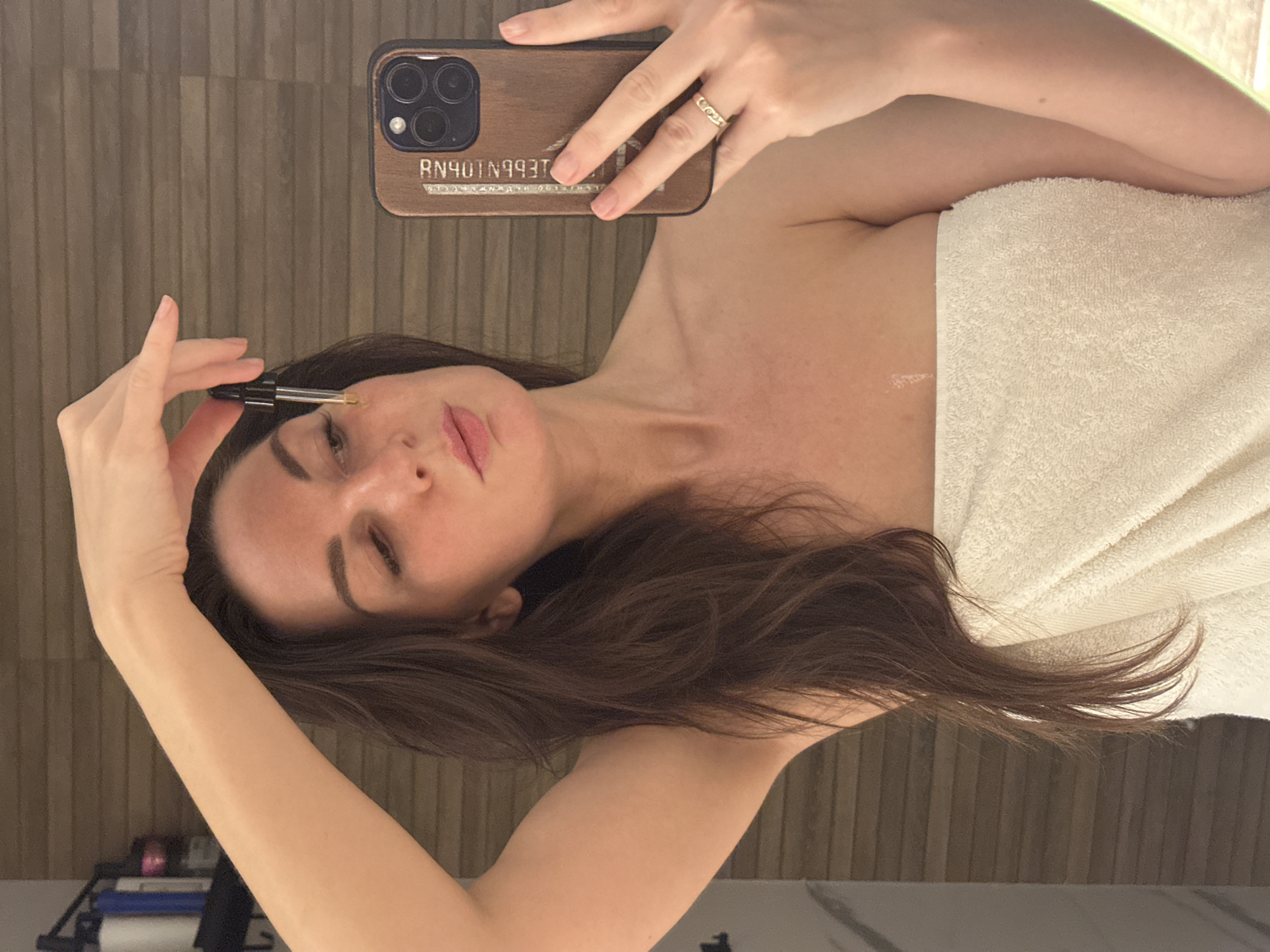 A fair-skinned woman with long brown hair is applying a liquid from a dropper onto her face while holding a smartphone in her right hand. She appears to be partially covered by a white towel. The smartphone has a wooden-looking case with a visible set of three camera lenses and an inscription, possibly "RN401N993T". The background consists of vertical wooden paneling. Objects are seen on a surface near the bottom of the image. The location is not clearly recognizable as Chaikovsky, Russia from the details provided.