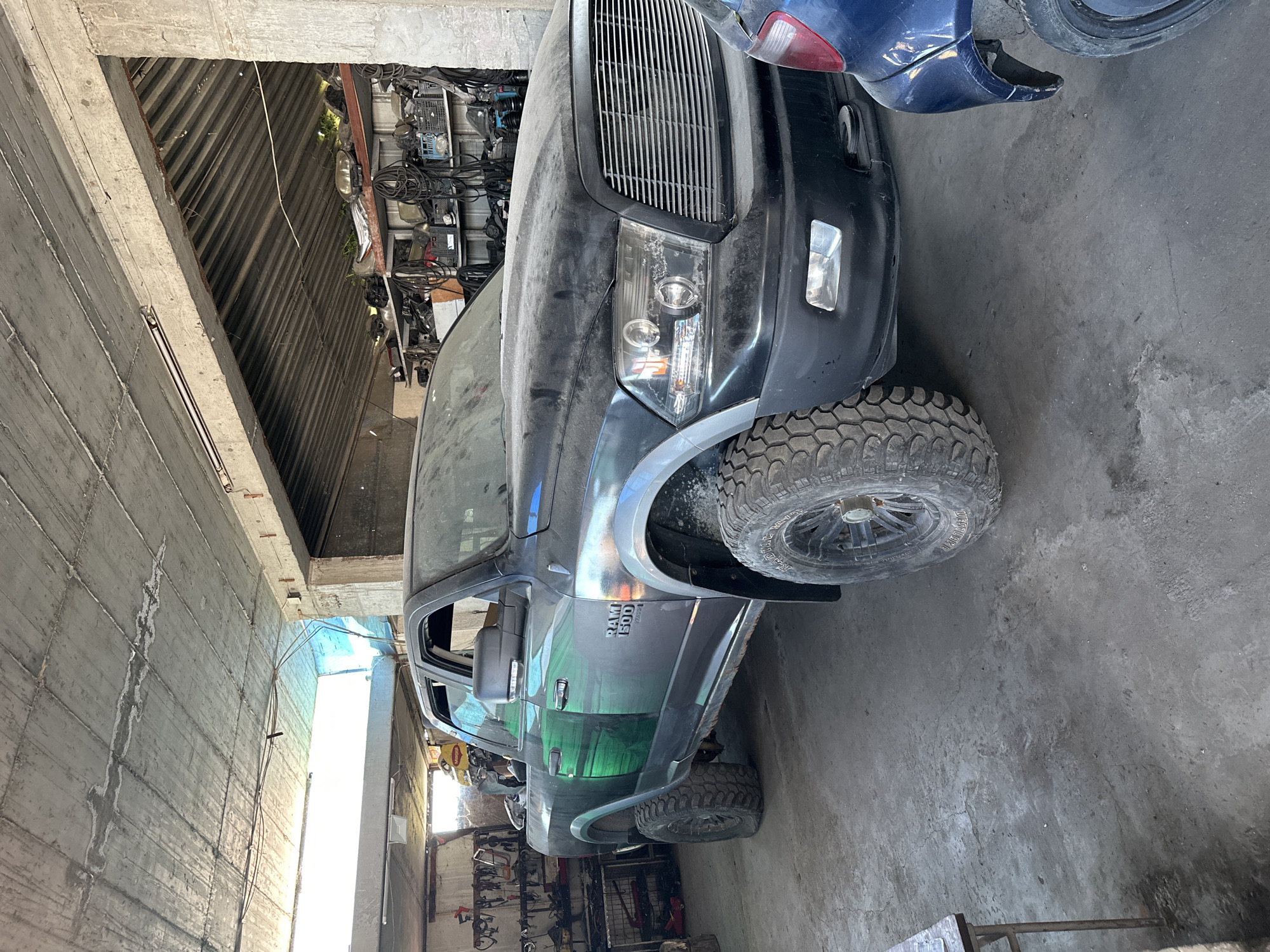 A dusty, black Dodge Ram 1500 pickup truck is parked indoors, possibly in a garage or workshop. The truck features large, off-road tires and has a custom green graphic design on its driver's side. The interior of the space is utilitarian, with exposed concrete ceilings and walls, and shelves stocked with car parts in the background. A blue car is partially visible to the right of the truck. The lighting suggests it is daytime, with natural light entering from an open area.